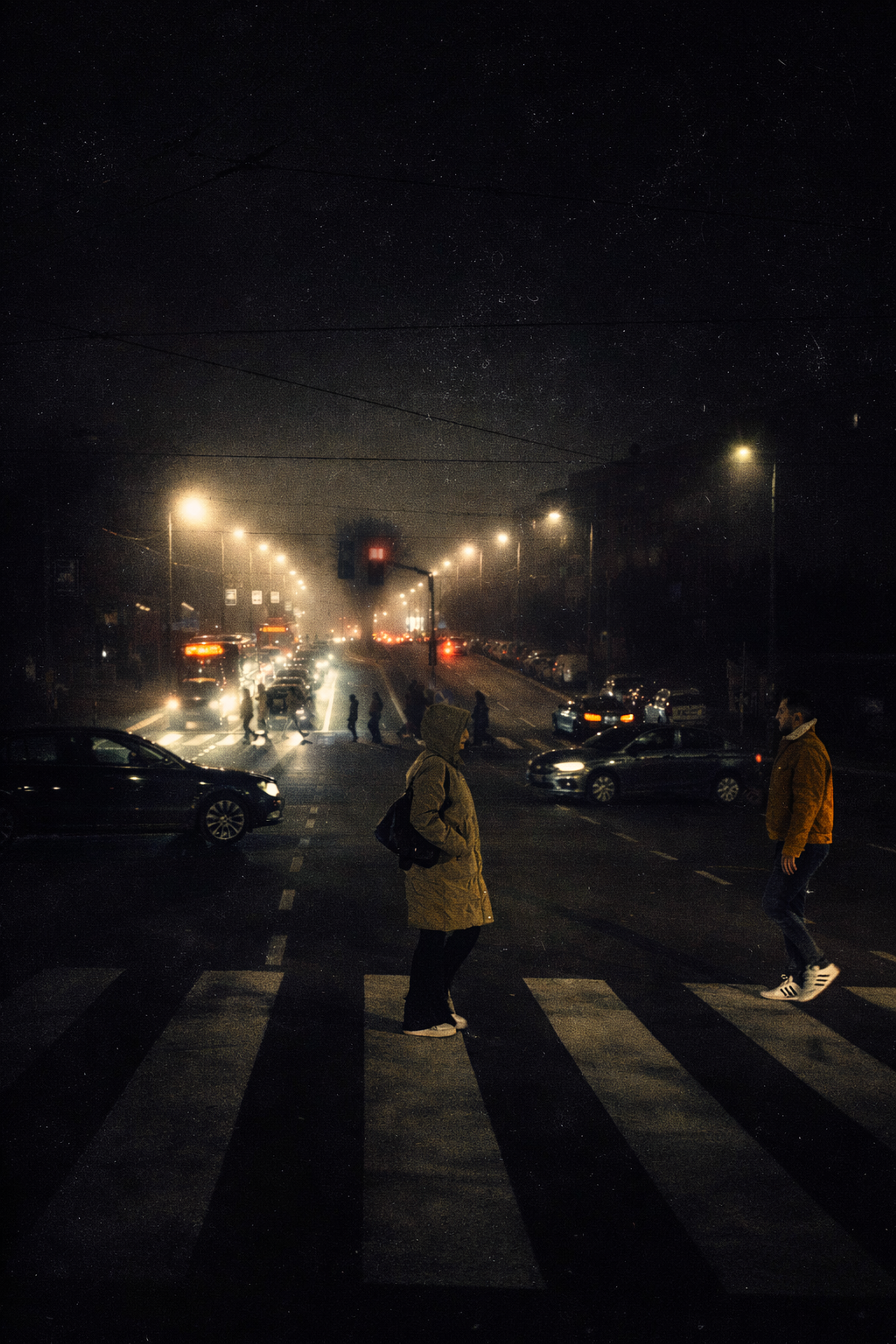 Atmospheric night street scene showing pedestrians crossing a marked zebra crosswalk under glowing streetlights and faint traffic beams.