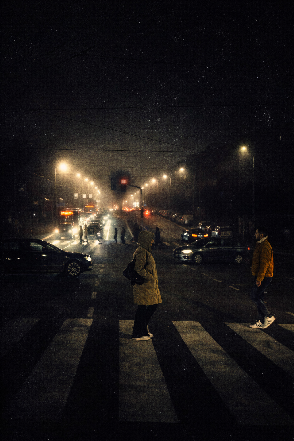 Atmospheric night street scene showing pedestrians crossing a marked zebra crosswalk under glowing streetlights and faint traffic beams.