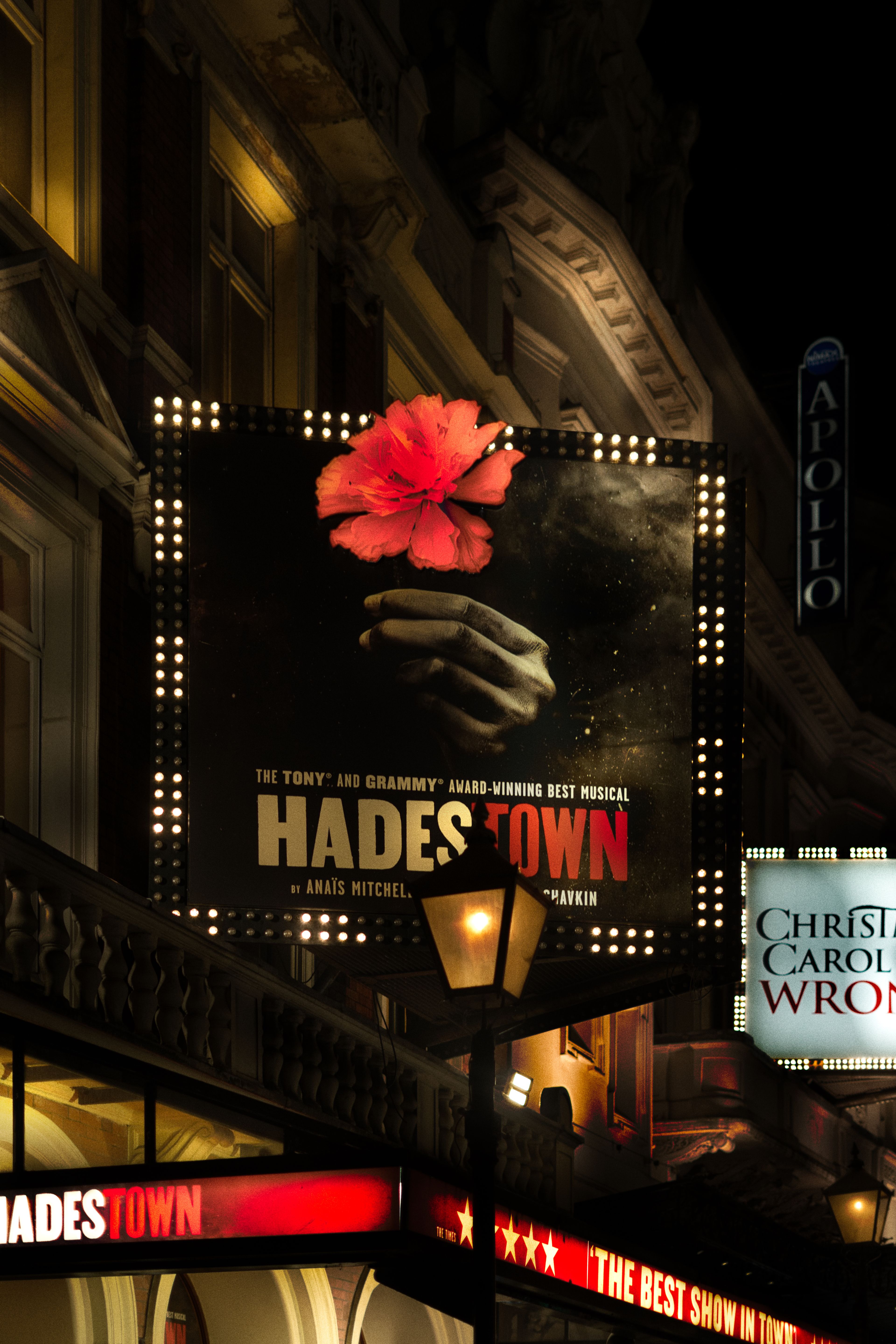 Low-angle night photograph of the Hadestown musical marquee displaying a hand holding a red flower above the illuminated theater entrance.