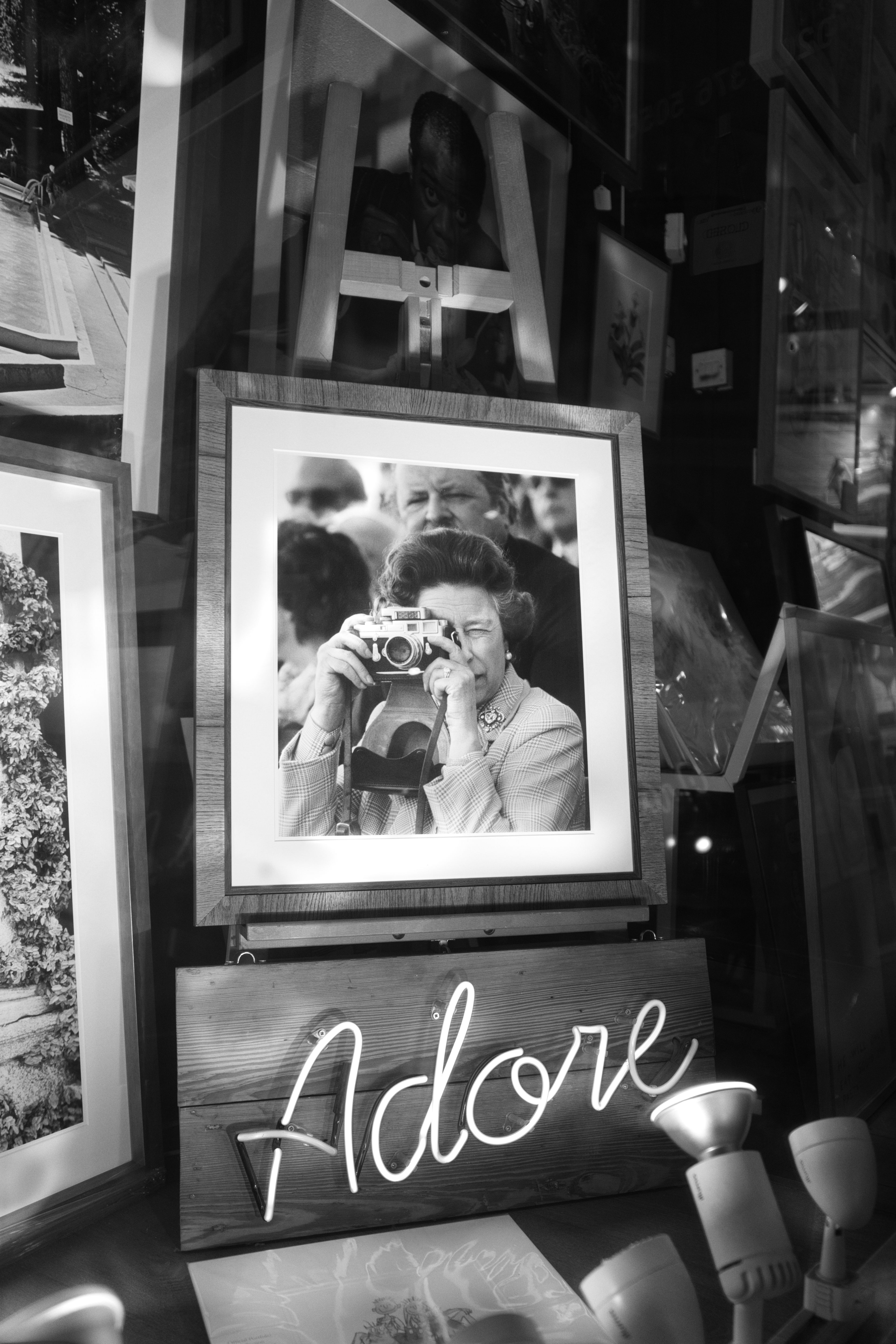 Black-and-white street photography showing a framed display with a vintage photograph of someone holding a camera, alongside a neon "Adore" sign