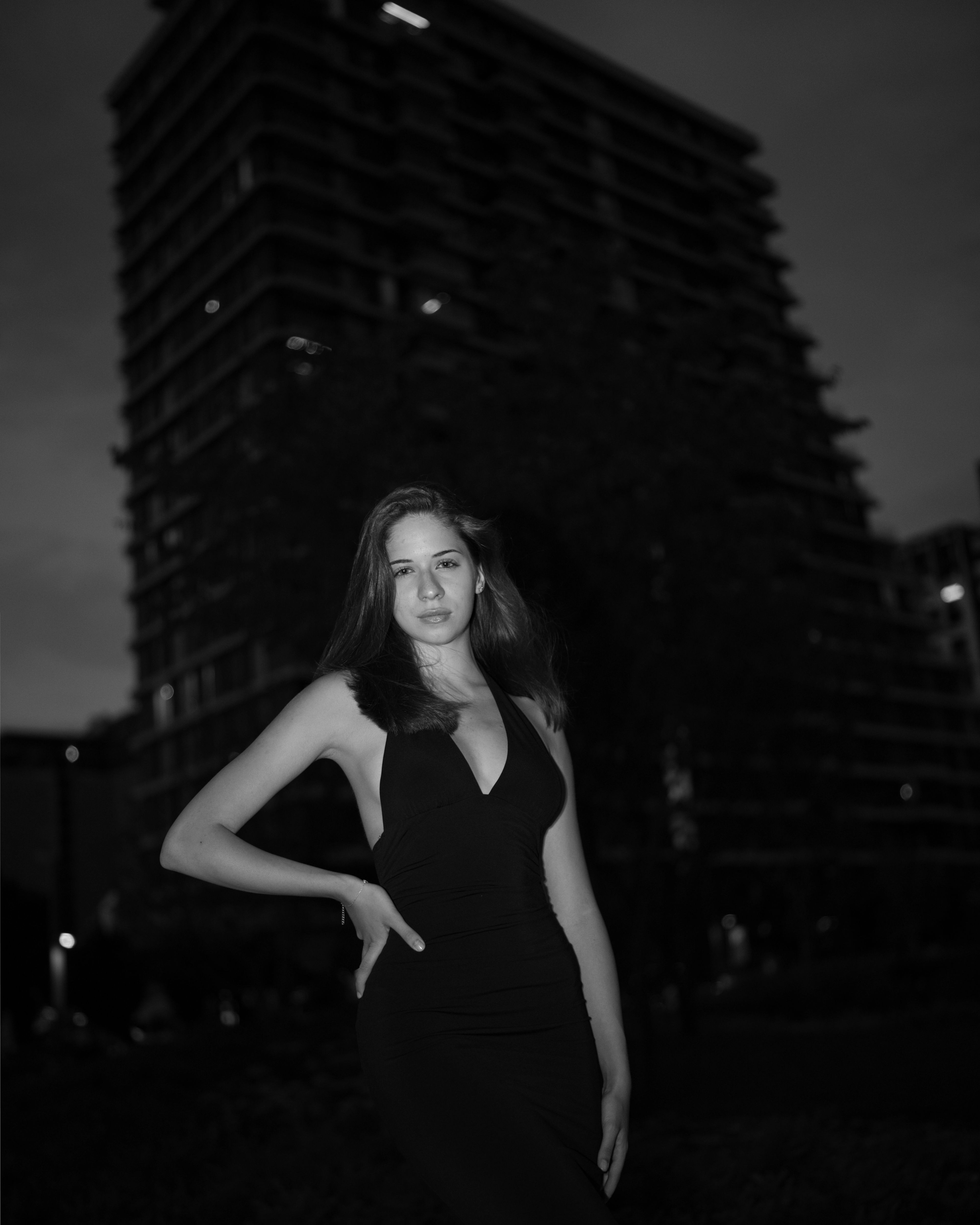 High-contrast monochrome portrait of a woman in a black dress posing outdoors against a towering brutalist building at night.