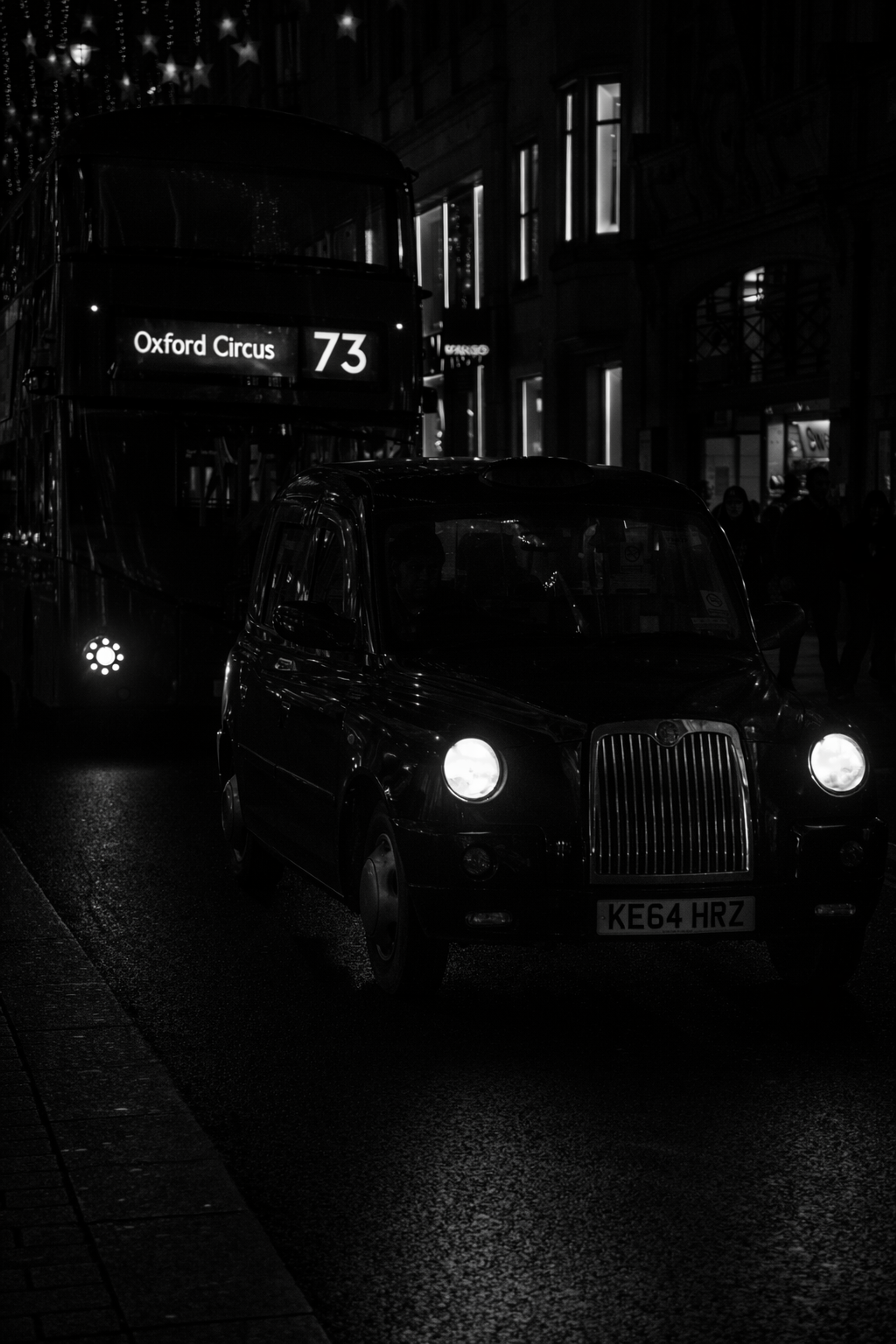 Moody black-and-white street photograph of a dark London taxi cab and a route 73 double-decker bus on a wet night street.