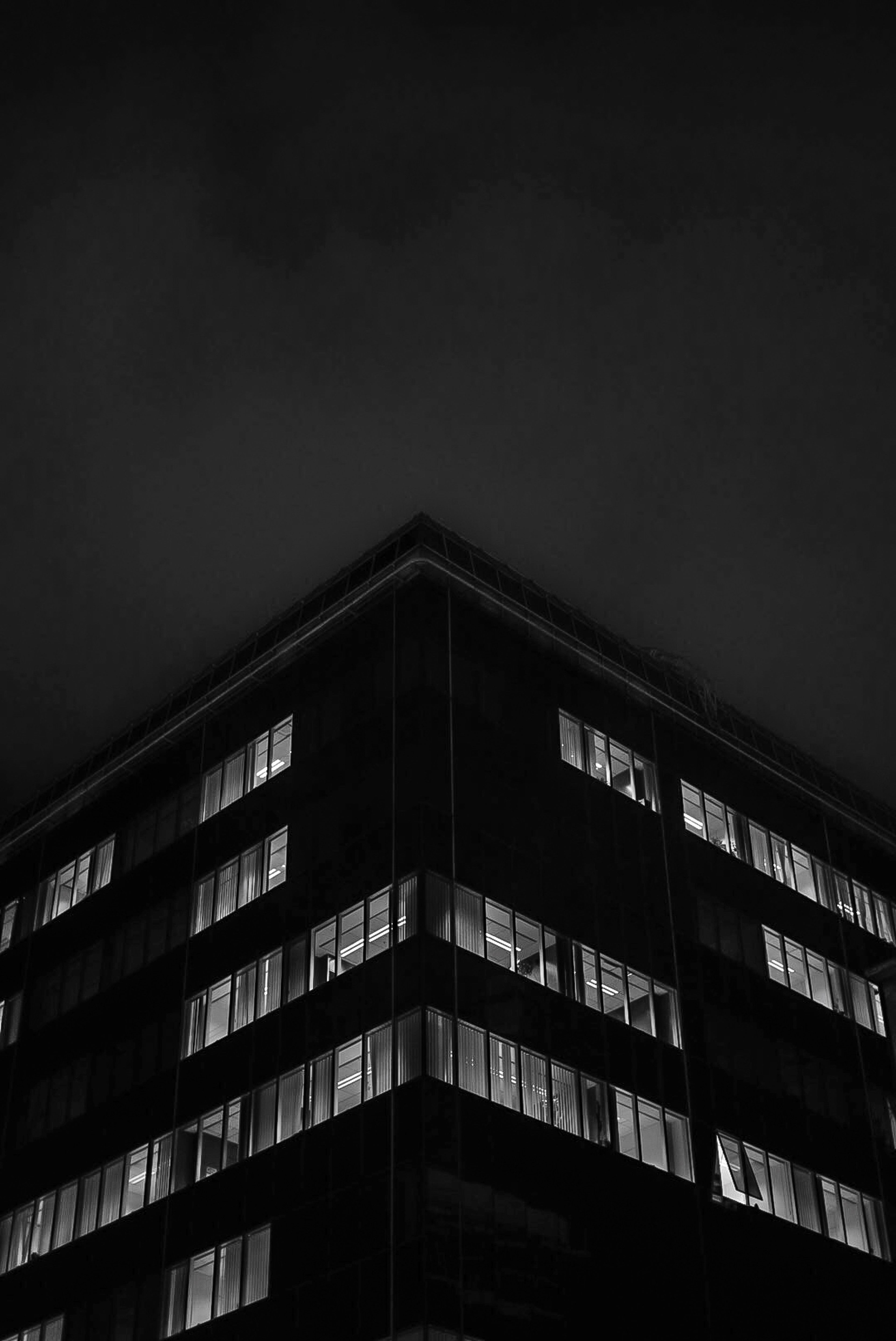 Low-angle black and white photograph of a modern glass skyscraper illuminated at night against a dark, moody sky.