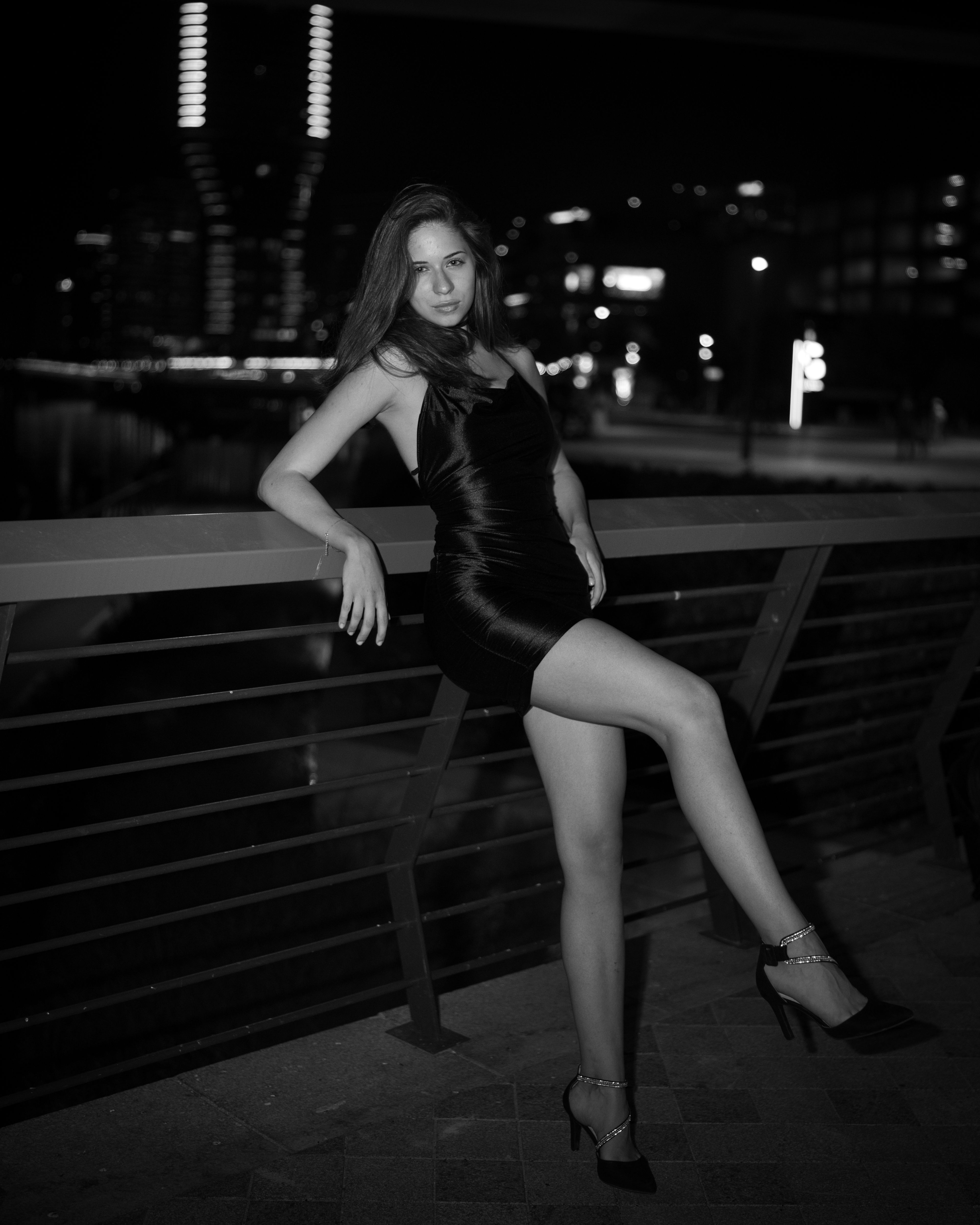Monochrome portrait of a woman leaning against an overpass railing with structural architecture and night lights behind her.