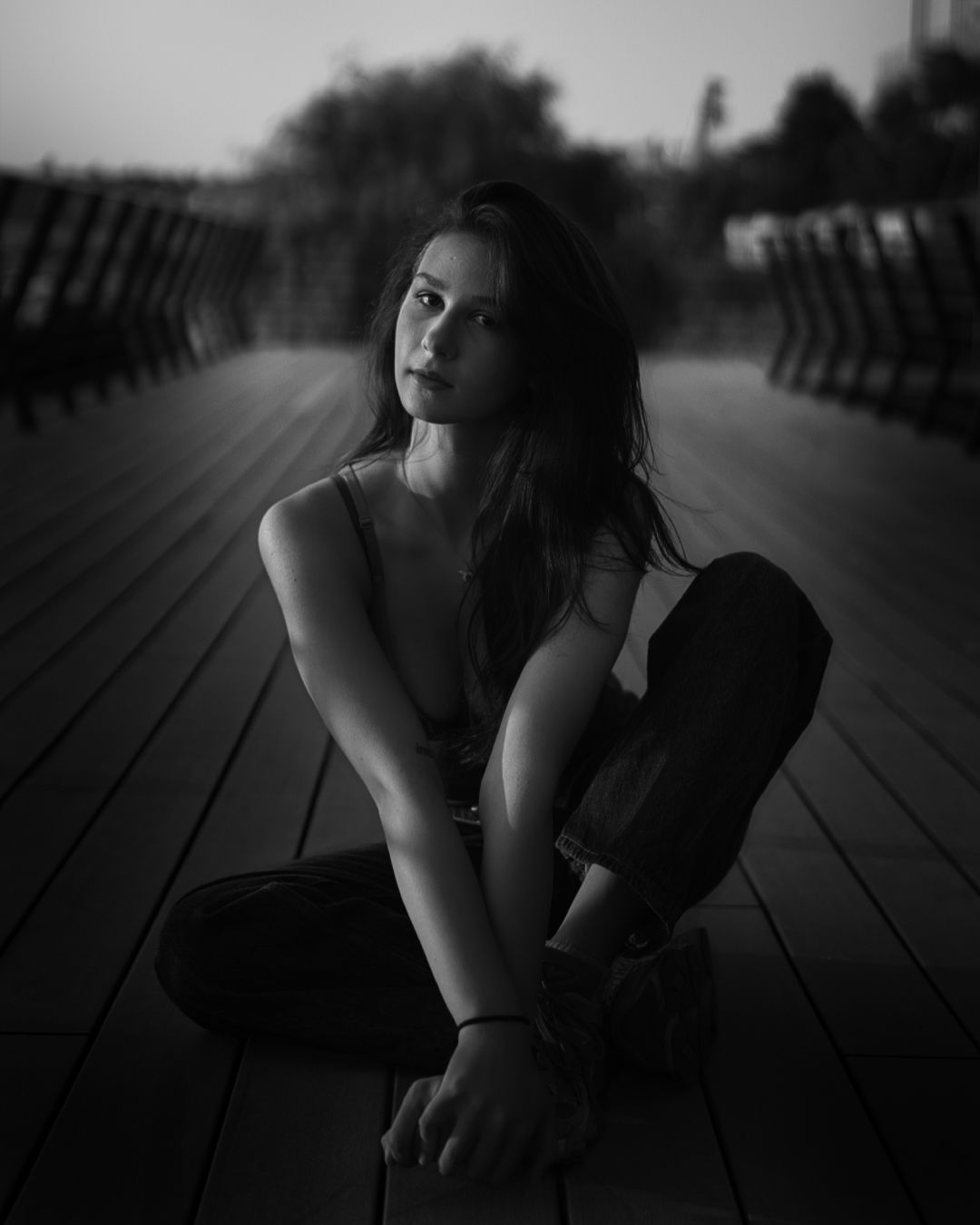 Atmospheric black-and-white portrait of a woman sitting gracefully on a wooden boardwalk at dusk.
