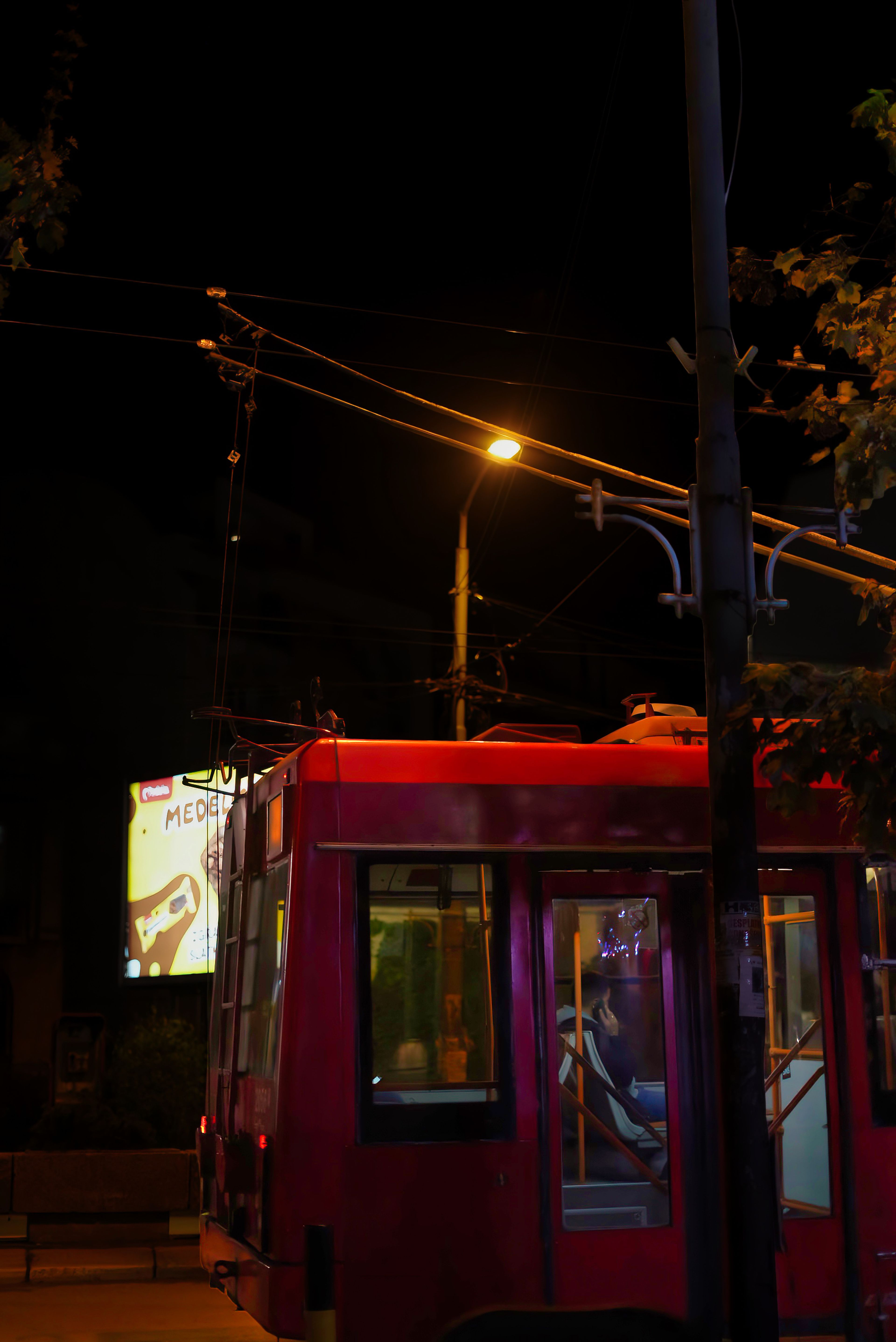 Vertical night shot of a red city tram moving under glowing streetlights and overhead power lines.