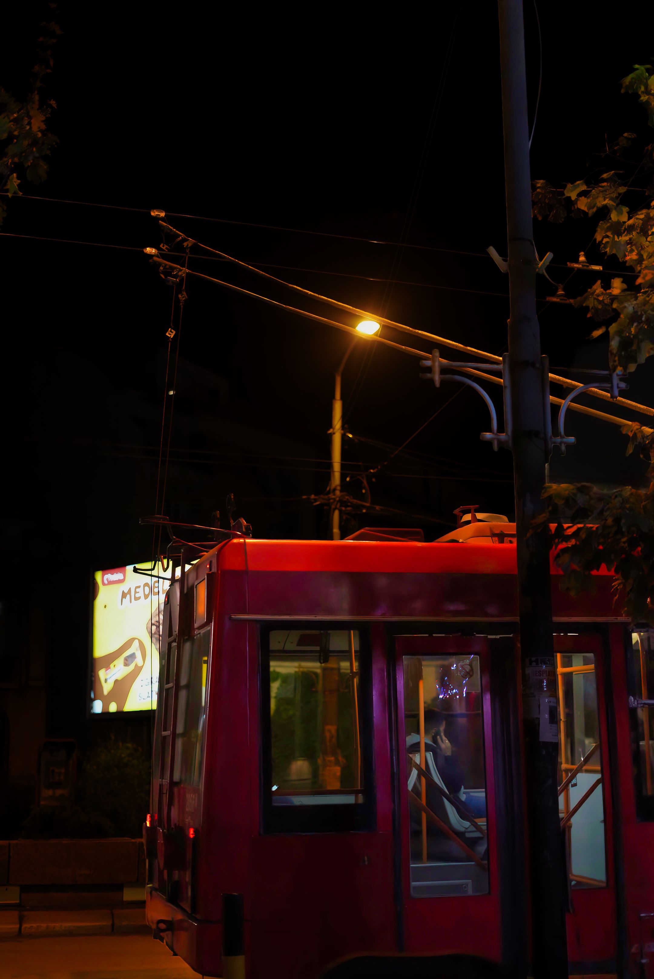 Vertical night shot of a red city tram moving under glowing streetlights and overhead power lines.