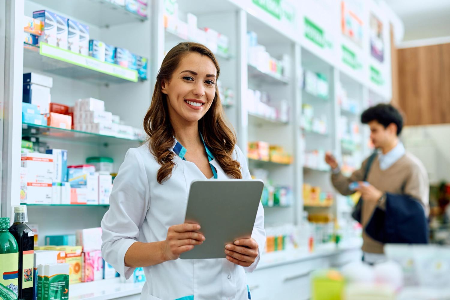 A female pharmacist smiling and holding an ipad, with a shelf of medicine behind her.