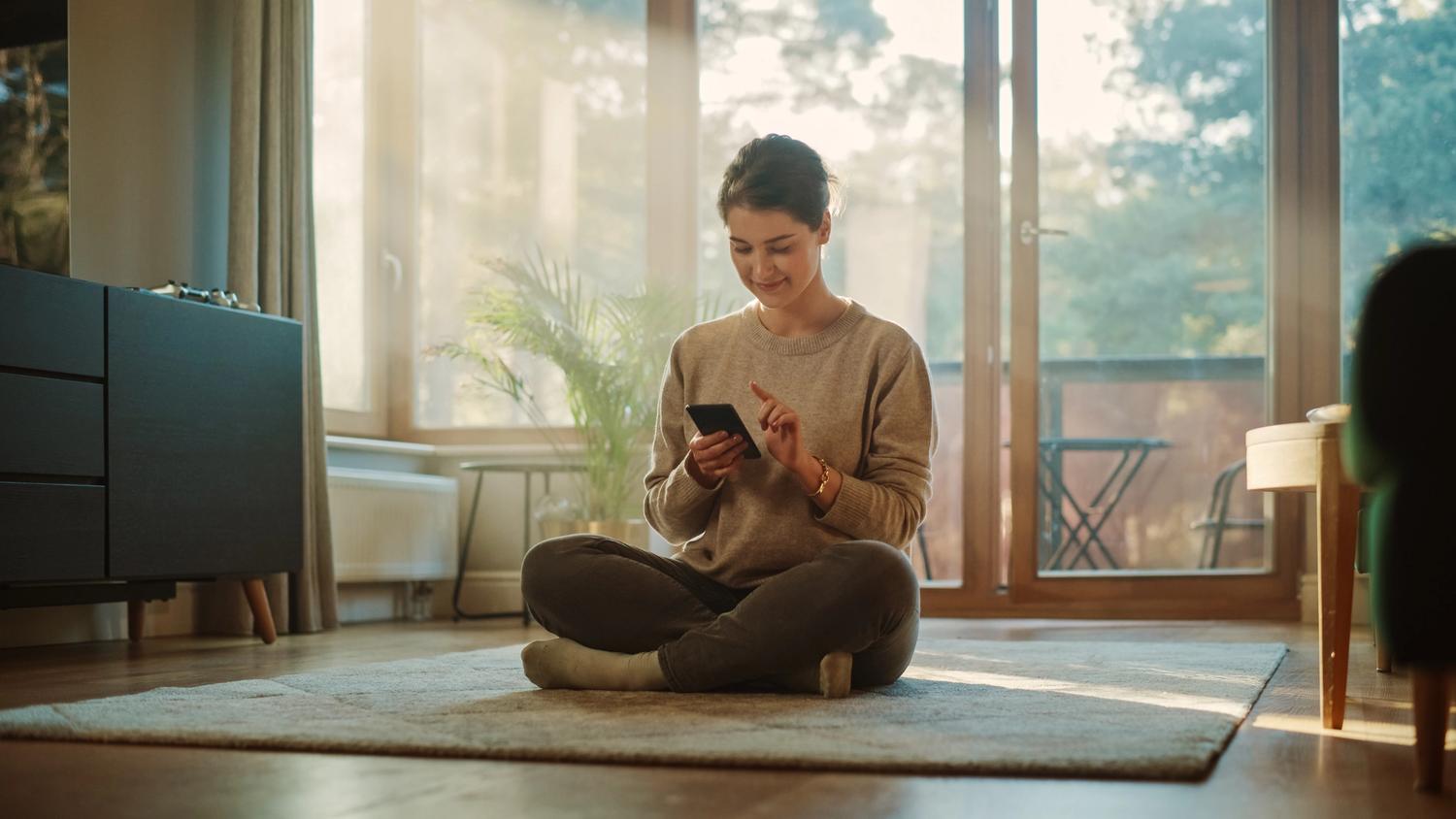 Woman sits crosslegged in lounge room smiling at phone