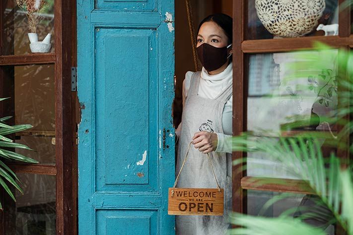 Lady in a mask opening a door and holding a sign saying 'WELCOME, WE ARE OPEN'