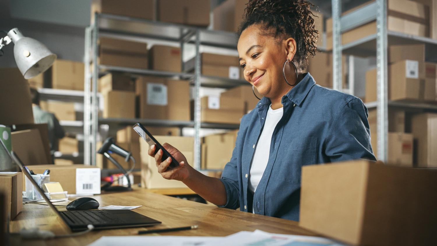 Small business owner smiles at phone at desk in warehouse 