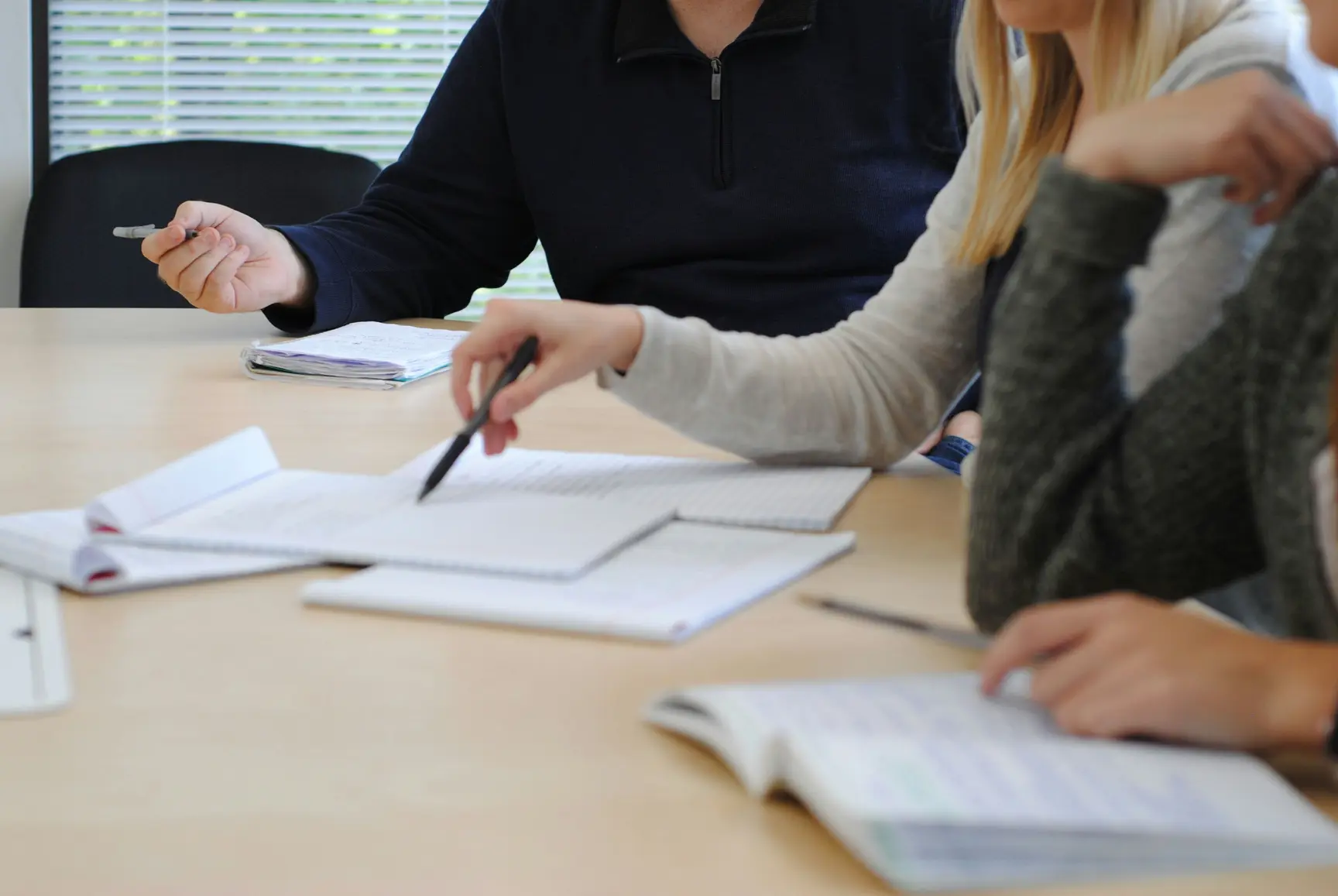 Claude responded: Three people reviewing documents and notebooks at a meeting table in a bright office environment.Three people reviewing documents and notebooks at a meeting table in a bright office environment.