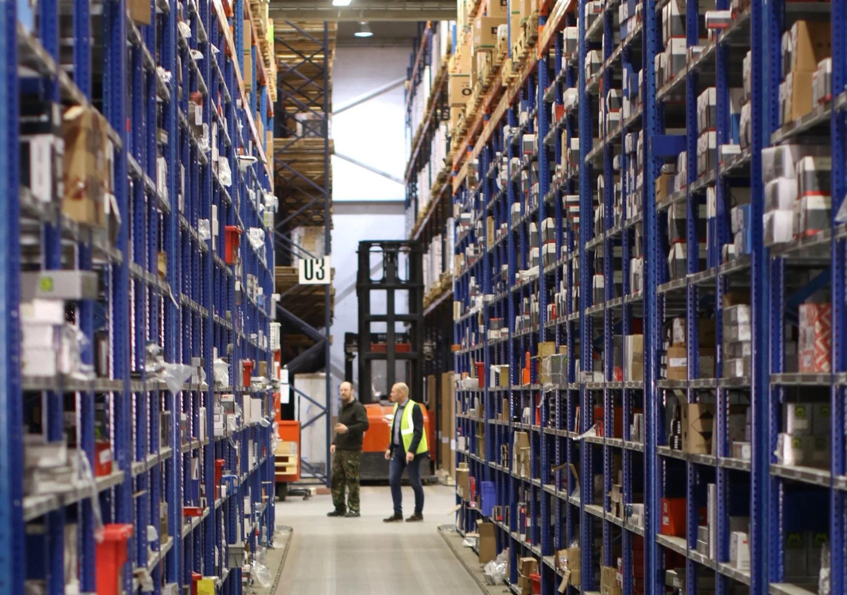 Two workers standing in a large warehouse aisle between tall blue shelving units filled with boxes, with a forklift visible.
