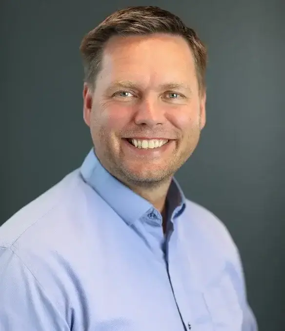 A man with short light brown hair and a beard, wearing a light blue collared shirt, smiles at the camera against a plain dark background.