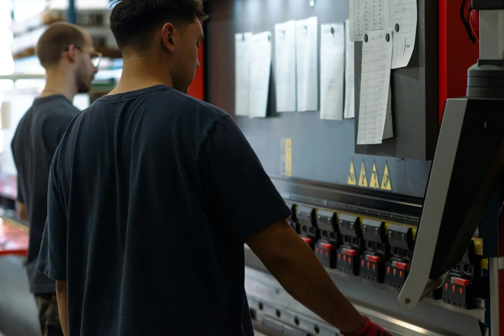 Claude responded: Two workers operating an industrial press brake machine on a factory floor with work instructions posted on the panel.Two workers operating an industrial press brake machine on a factory floor with work instructions posted on the panel.