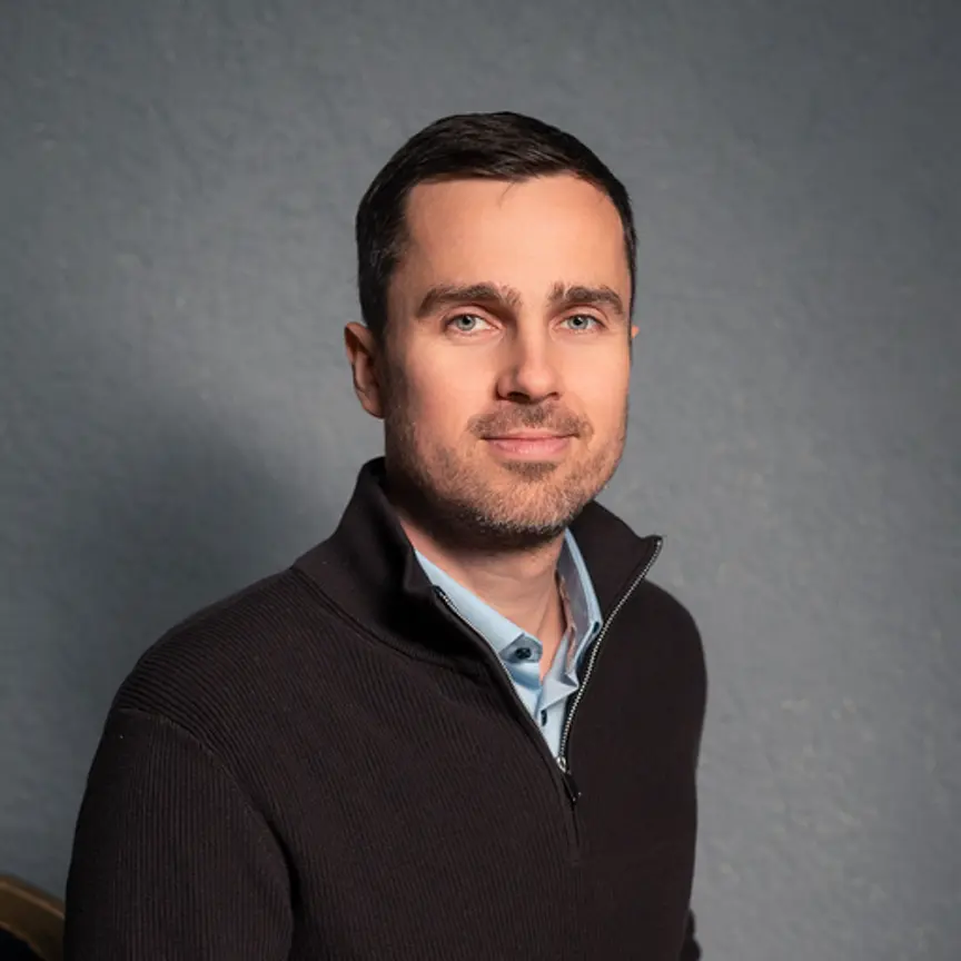 A man with short brown hair and a trimmed beard is wearing a dark zip-up sweater over a light blue collared shirt, posing in front of a plain gray textured background.