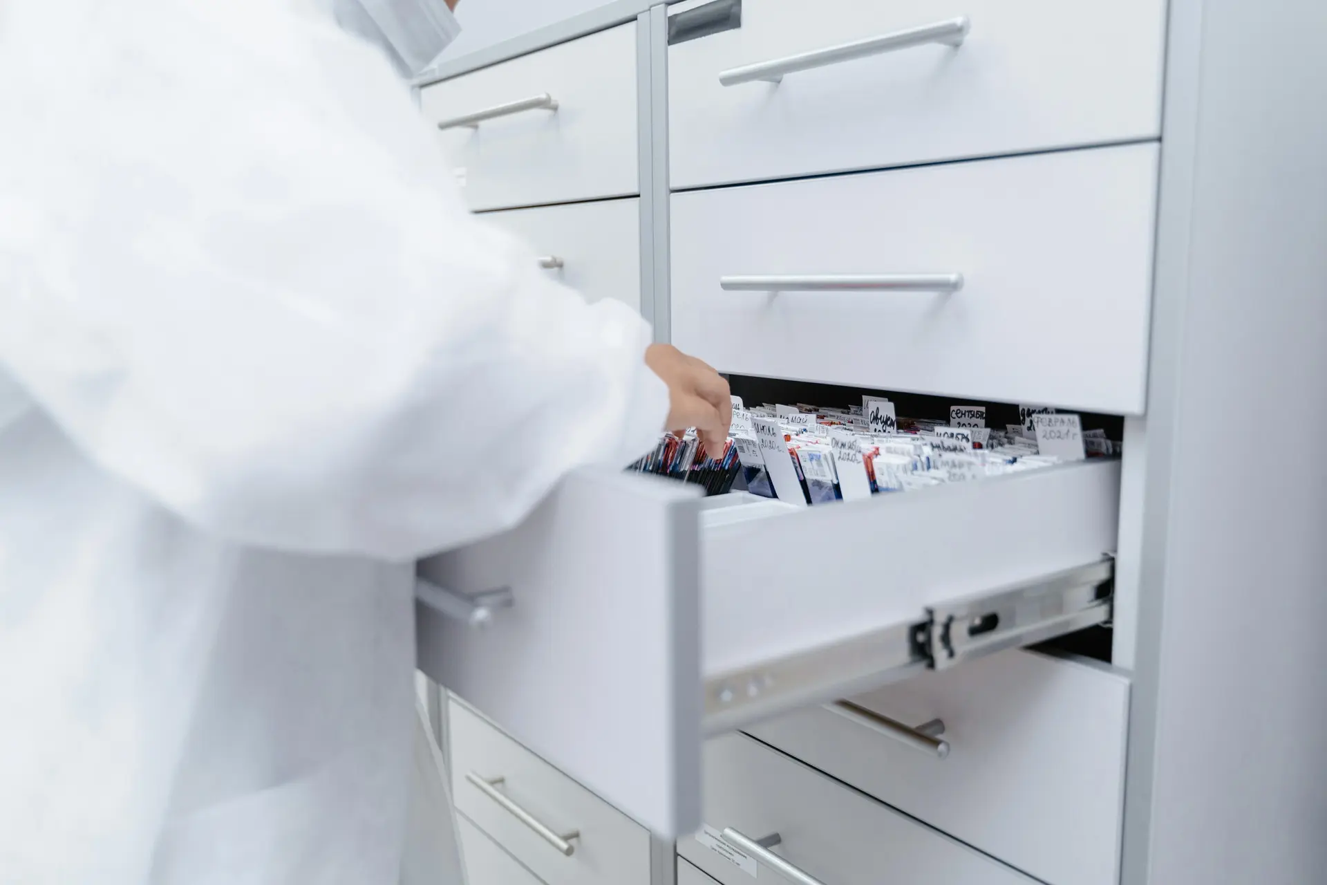 A person in a white lab coat retrieving labelled medication packages from an open drawer in a pharmacy storage cabinet.