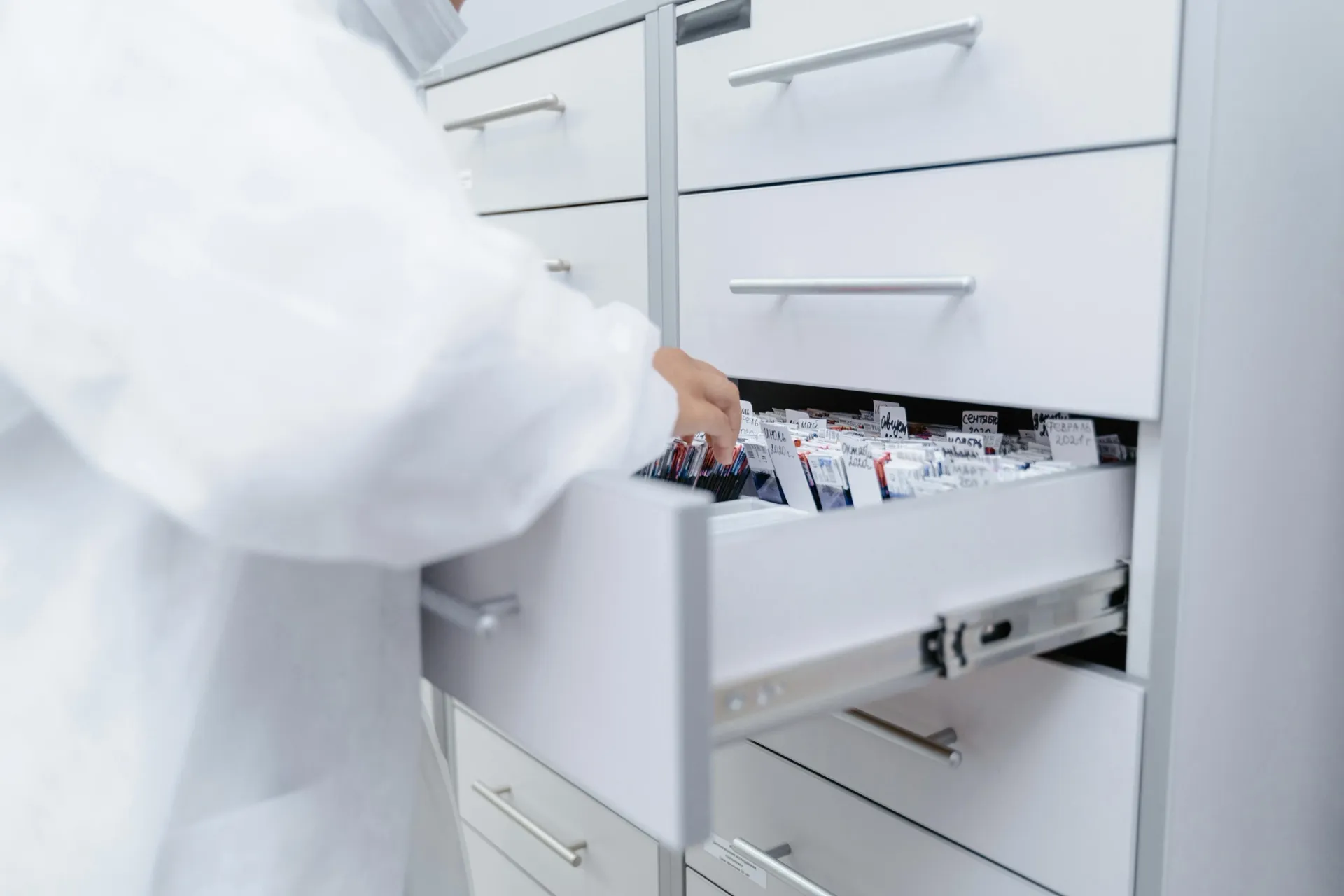 A person in a white lab coat retrieving labelled medication packages from an open drawer in a pharmacy storage cabinet.