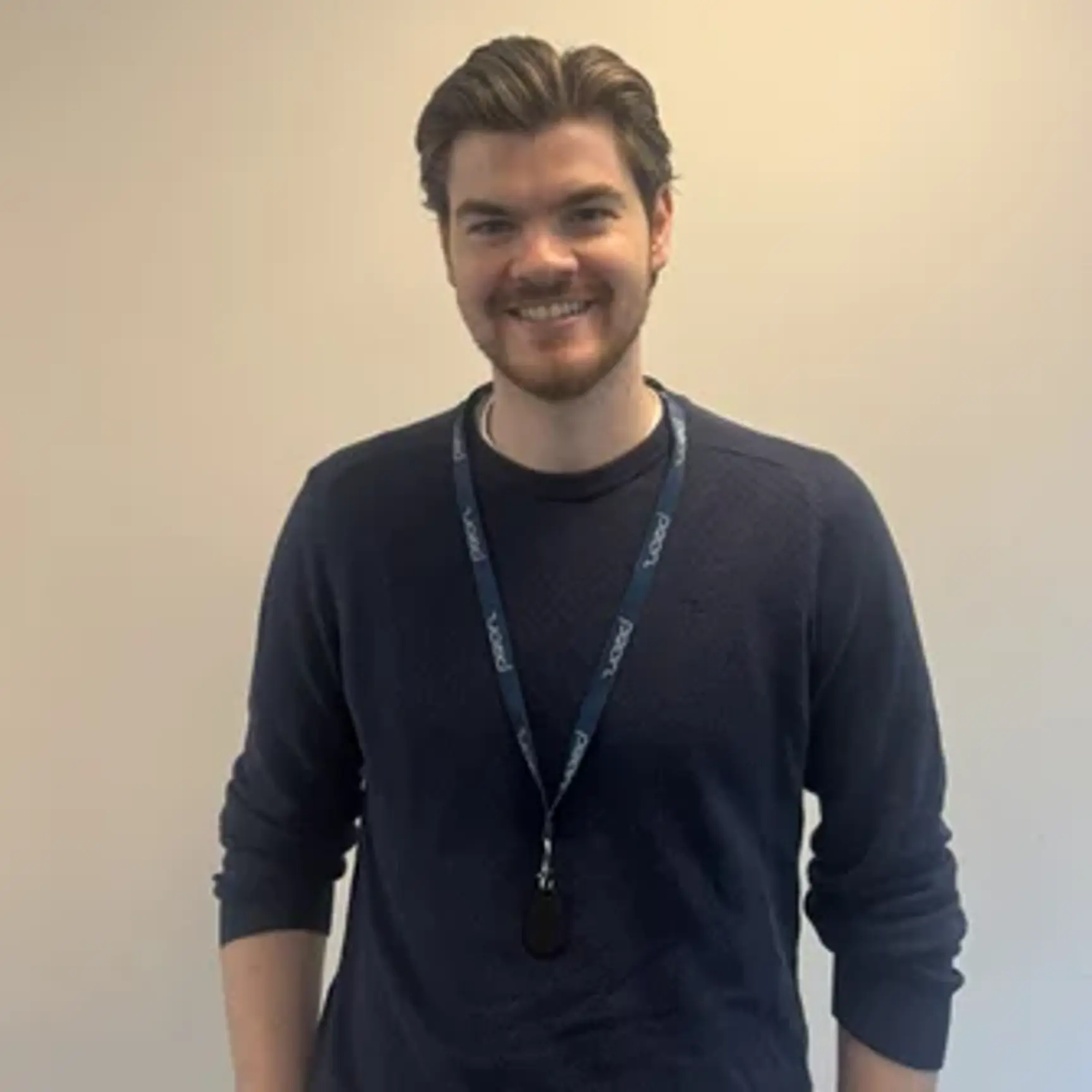 Portrait of a smiling man in a dark blue sweater and a lanyard, standing against a neutral, light wall.