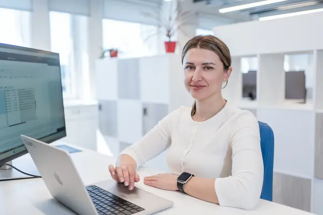 Woman in white shirt sitting and working by a desk with her laptop.