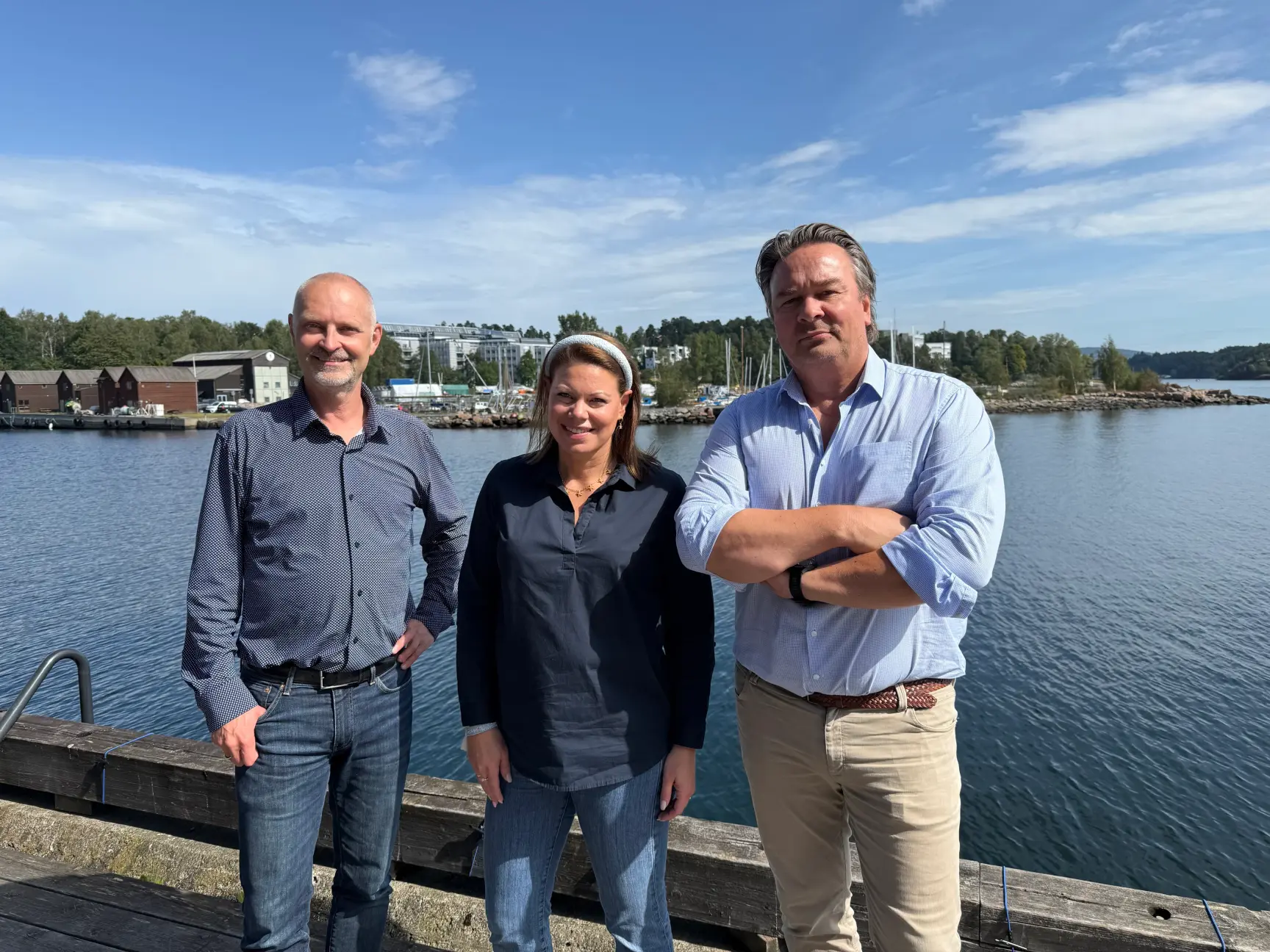 Three people stand smiling on a wooden pier by a blue fjord, with a shoreline and buildings in the background.