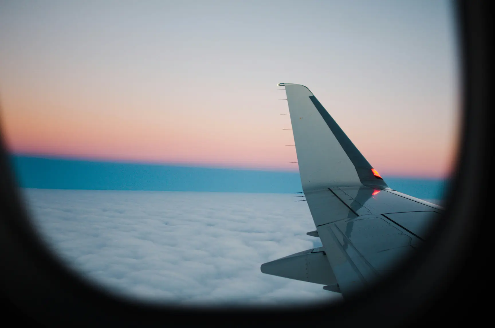 Aircraft wing viewed through a cabin window above a cloud layer with a pink and blue sunset horizon.Aircraft wing viewed through a cabin window above a cloud layer with a pink and blue sunset horizon.