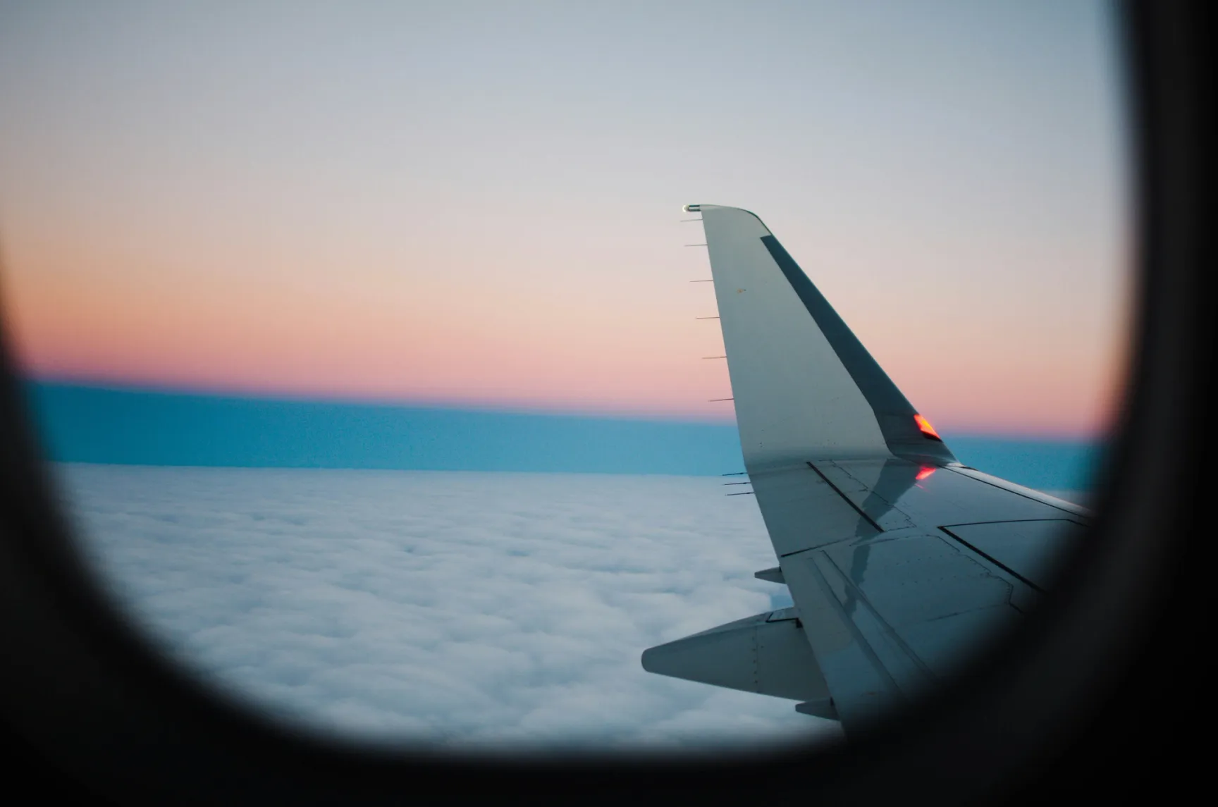 Aircraft wing viewed through a cabin window above a cloud layer with a pink and blue sunset horizon.Aircraft wing viewed through a cabin window above a cloud layer with a pink and blue sunset horizon.