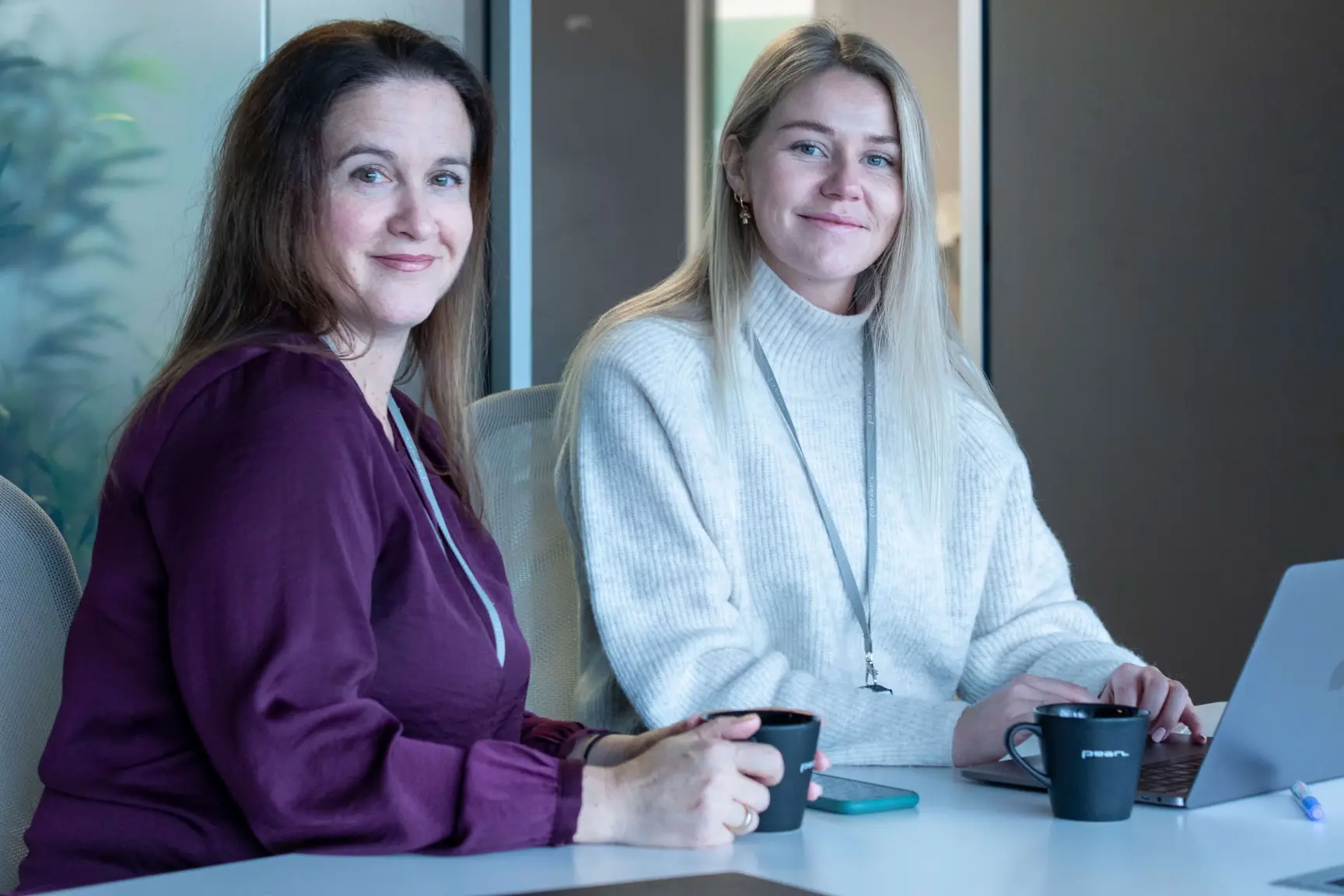 Two smiling women at a white office desk with coffee mugs and a laptop, wearing a purple blouse and a white sweater.