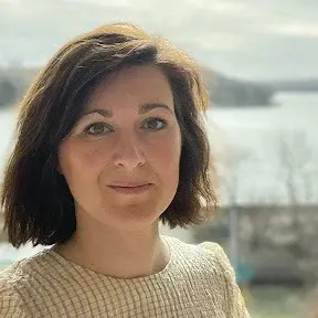 Portrait of a woman with brown hair and a checkered blouse, with a blurred outdoor landscape in the background.