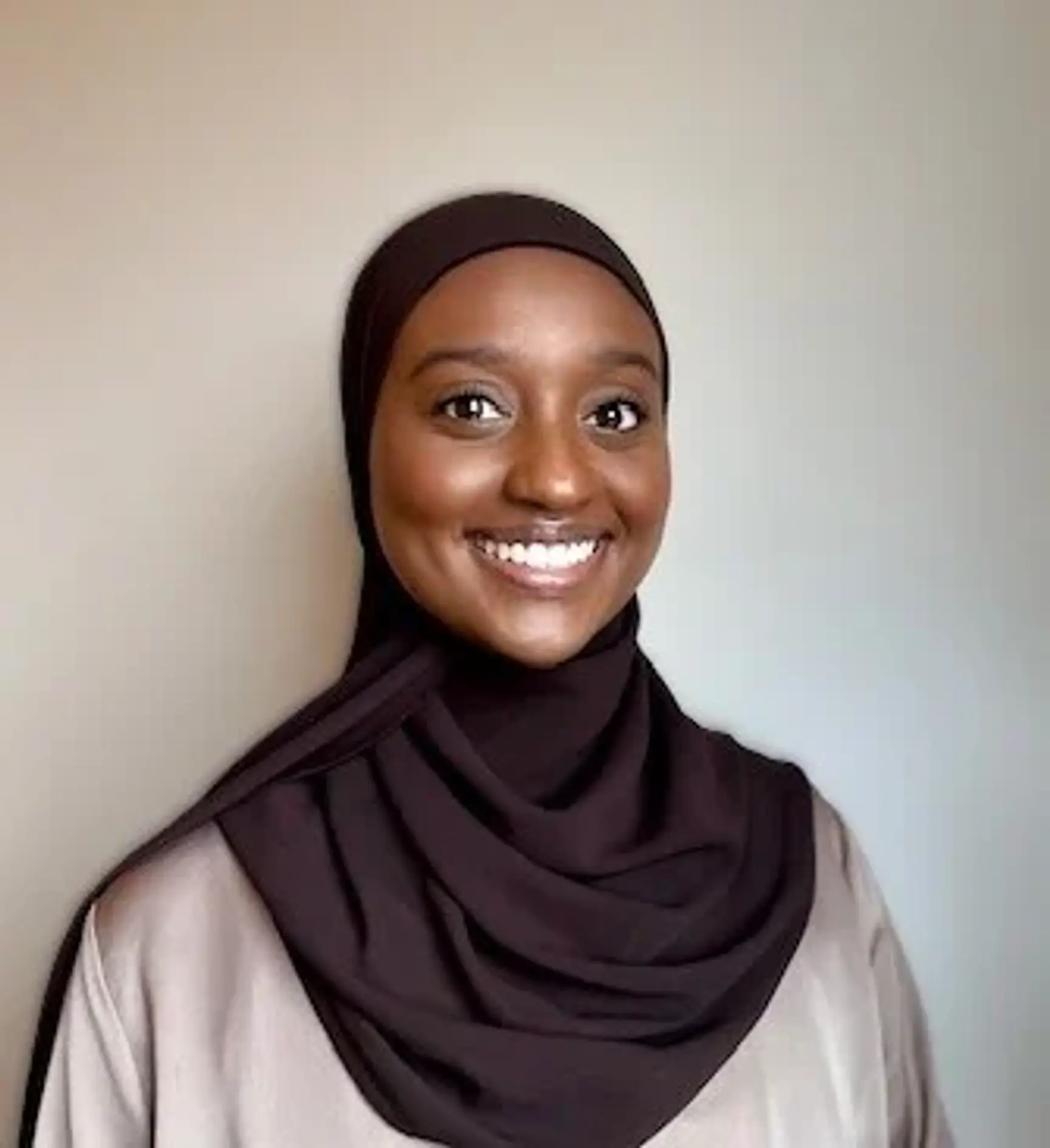 Portrait of a smiling woman wearing a dark brown hijab and beige top against a neutral, light background.