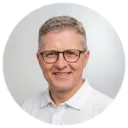 Smiling man with glasses in a white shirt photographed against a light grey background in a circular crop portrait photo.