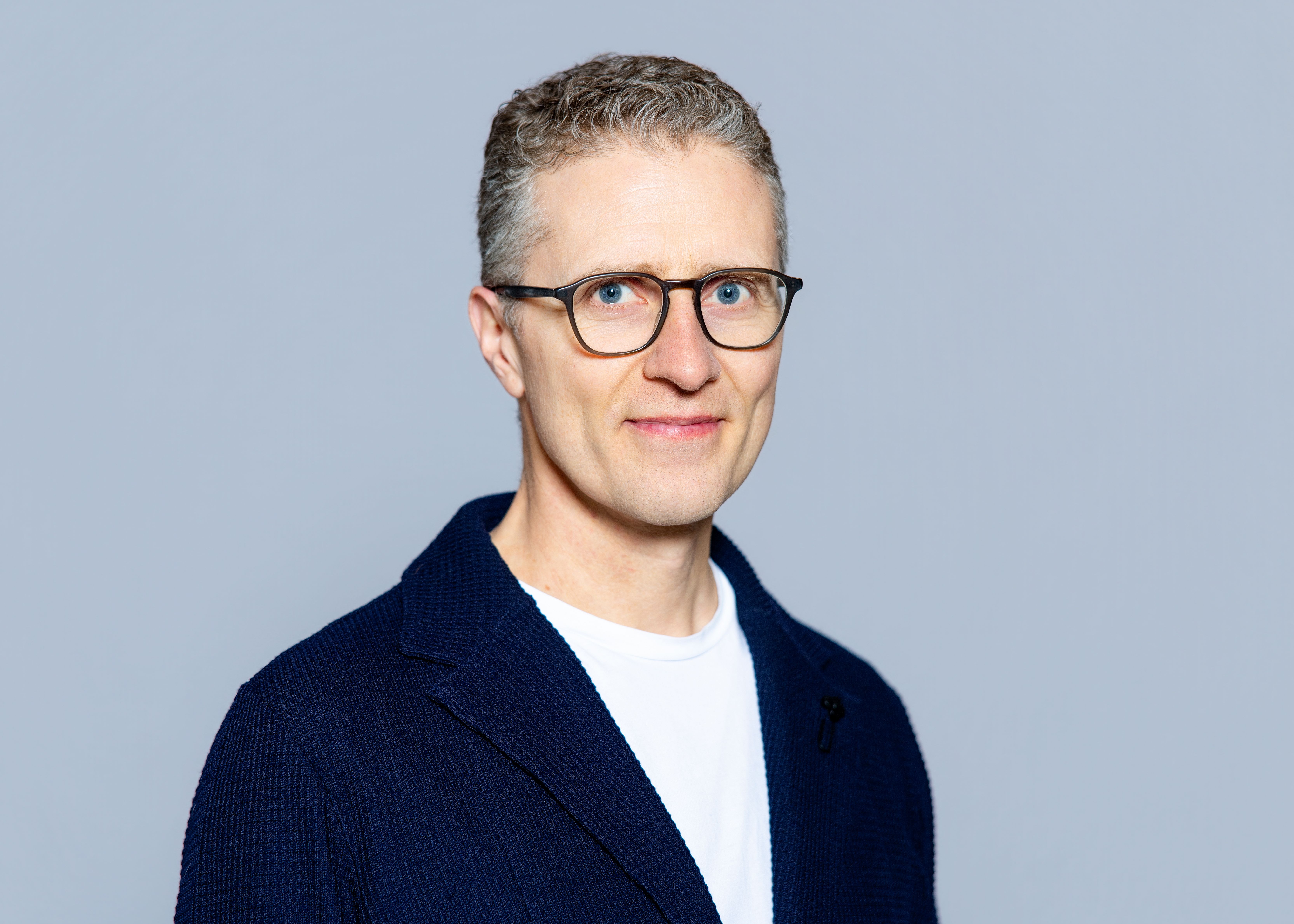 Smiling man with glasses in a white shirt photographed against a light grey background in a circular crop portrait photo.