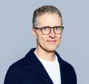 Smiling man with glasses in a white shirt photographed against a light grey background in a circular crop portrait photo.