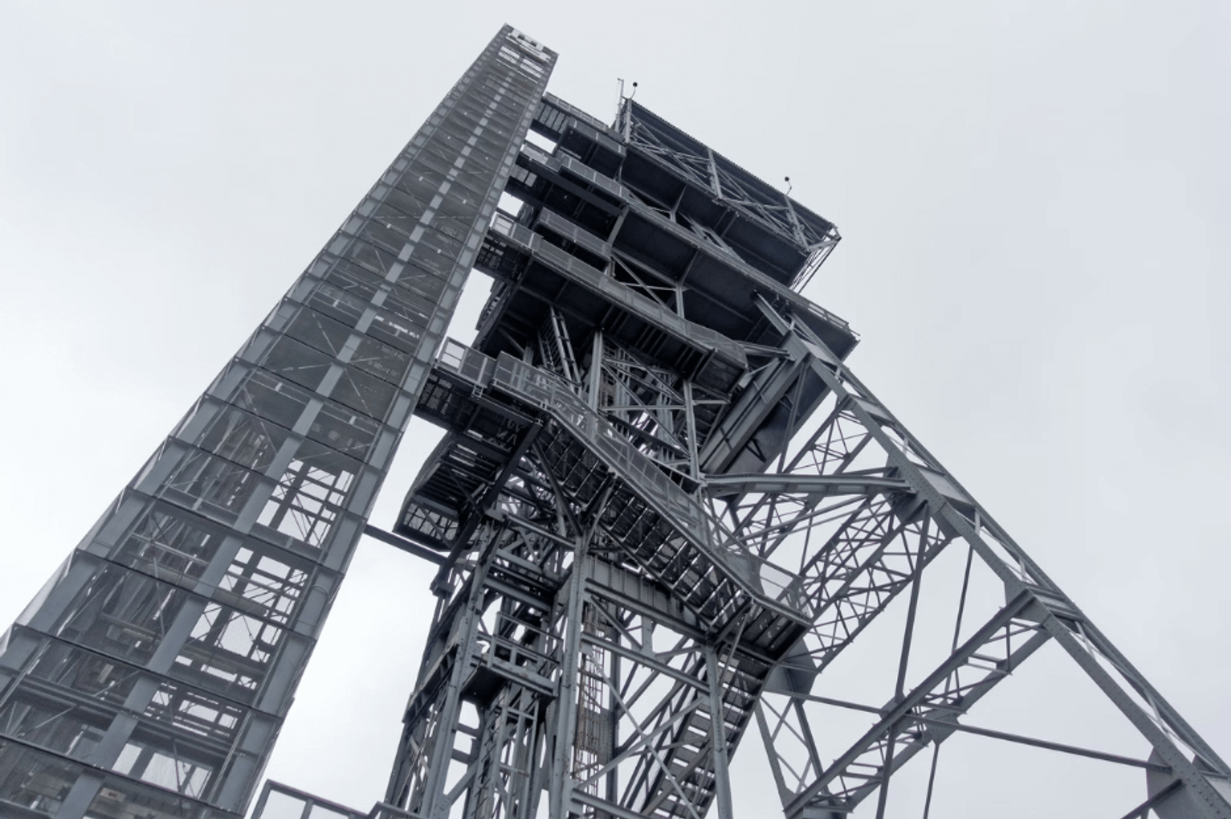 Low-angle view looking up at a large industrial steel framework tower and structure against an overcast grey sky.