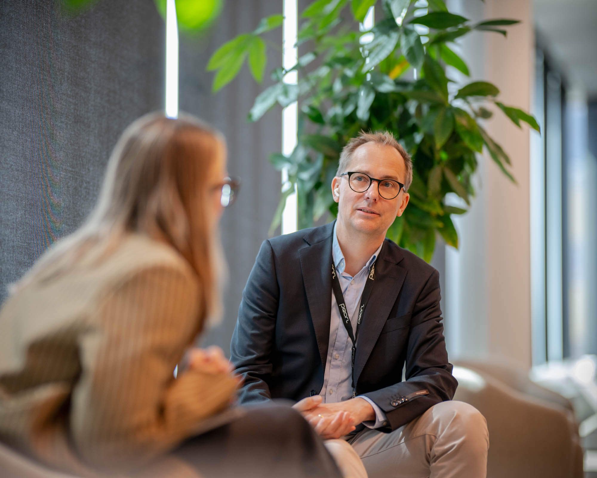 Man with glasses and a black jacket sits down in a conversation with a blonde woman.