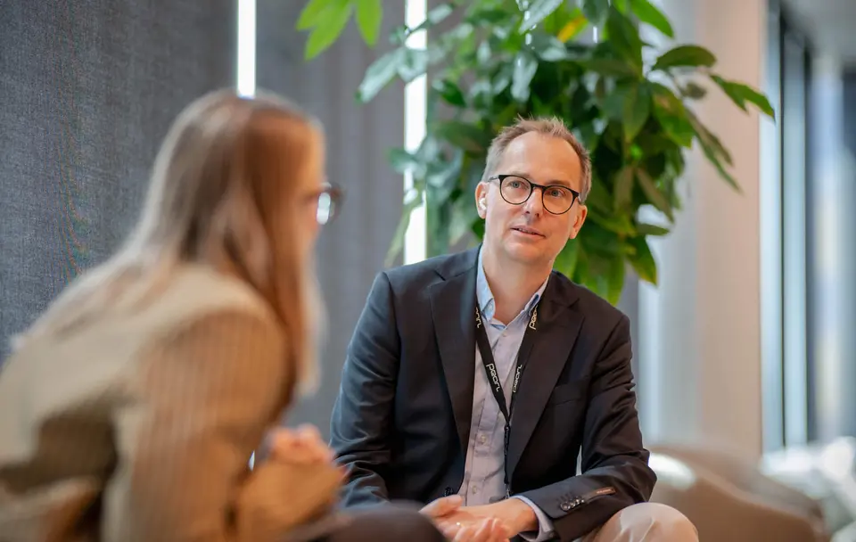 Man with glasses and a black jacket sits down in a conversation with a blonde woman.
