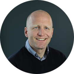 Smiling bald man in a dark sweater photographed against a dark grey background in a circular crop portrait photo.