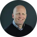 Smiling bald man in a dark sweater photographed against a dark grey background in a circular crop portrait photo.