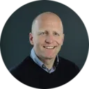 Smiling bald man in a dark sweater photographed against a dark grey background in a circular crop portrait photo.