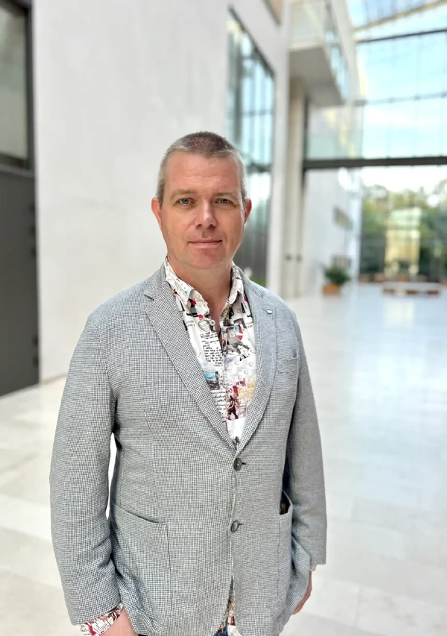 A man with short hair wearing a gray blazer and patterned shirt stands indoors in a bright, modern atrium with large windows and white walls.