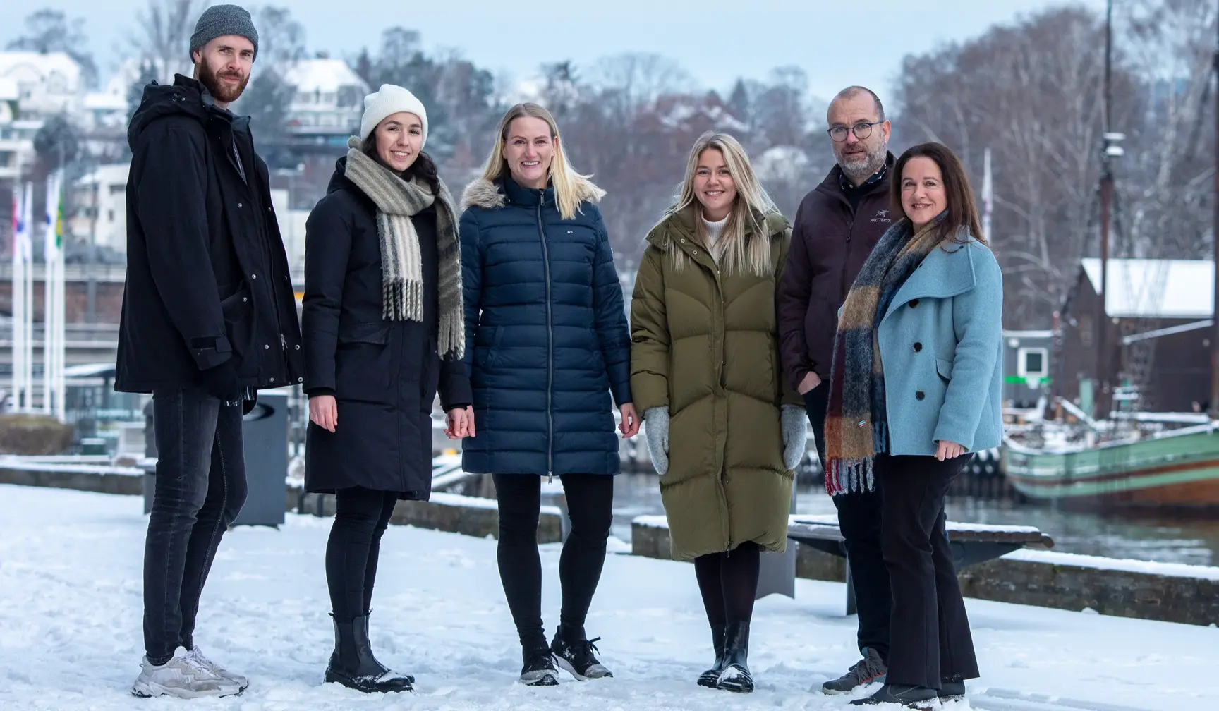 Six people standing in a group outdoors in a snowy landscape, wearing winter coats with water in the background.
