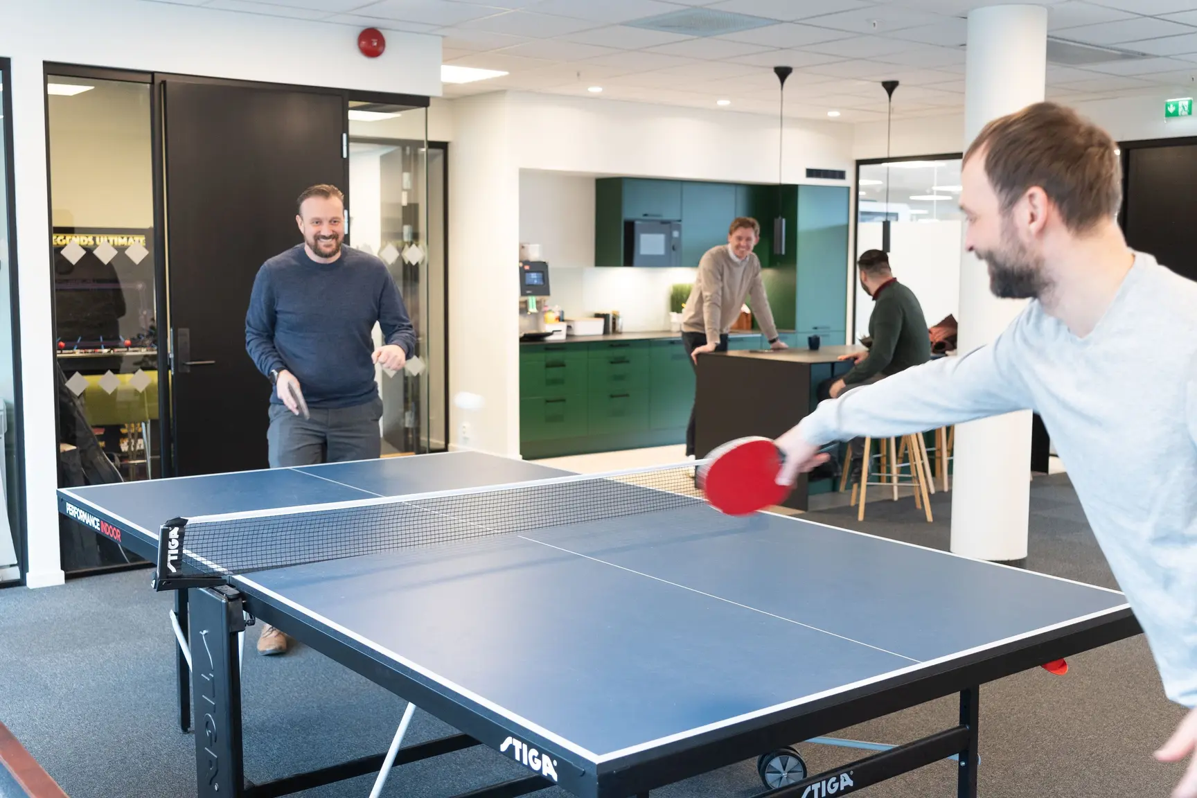 Two men playing table tennis in an open office space with colleagues and a green kitchen in the background.