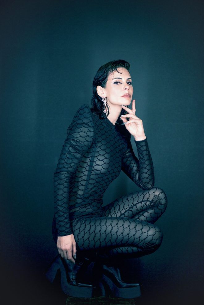 Rebekah crouching in a patterned black outfit and platform boots against a dark teal backdrop