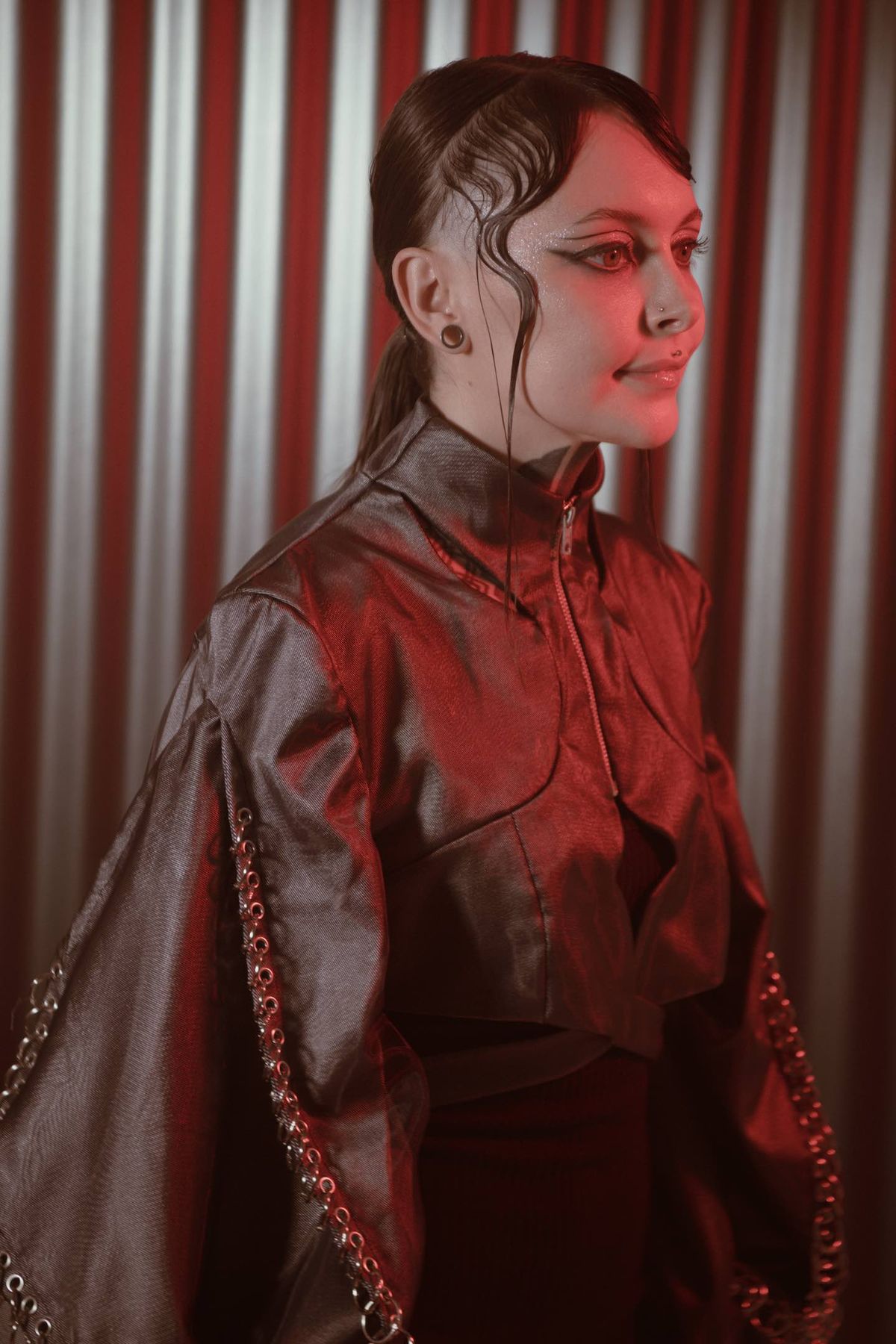Krista in profile wearing a metallic cropped jacket with chain trim against a red-lit metal wall