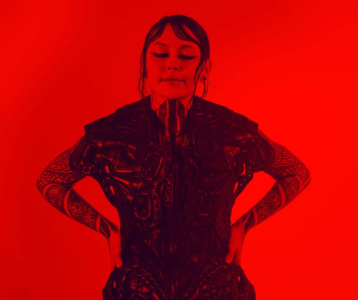Krista with eyes closed and hands on hips, wearing a sculptural black top under red light