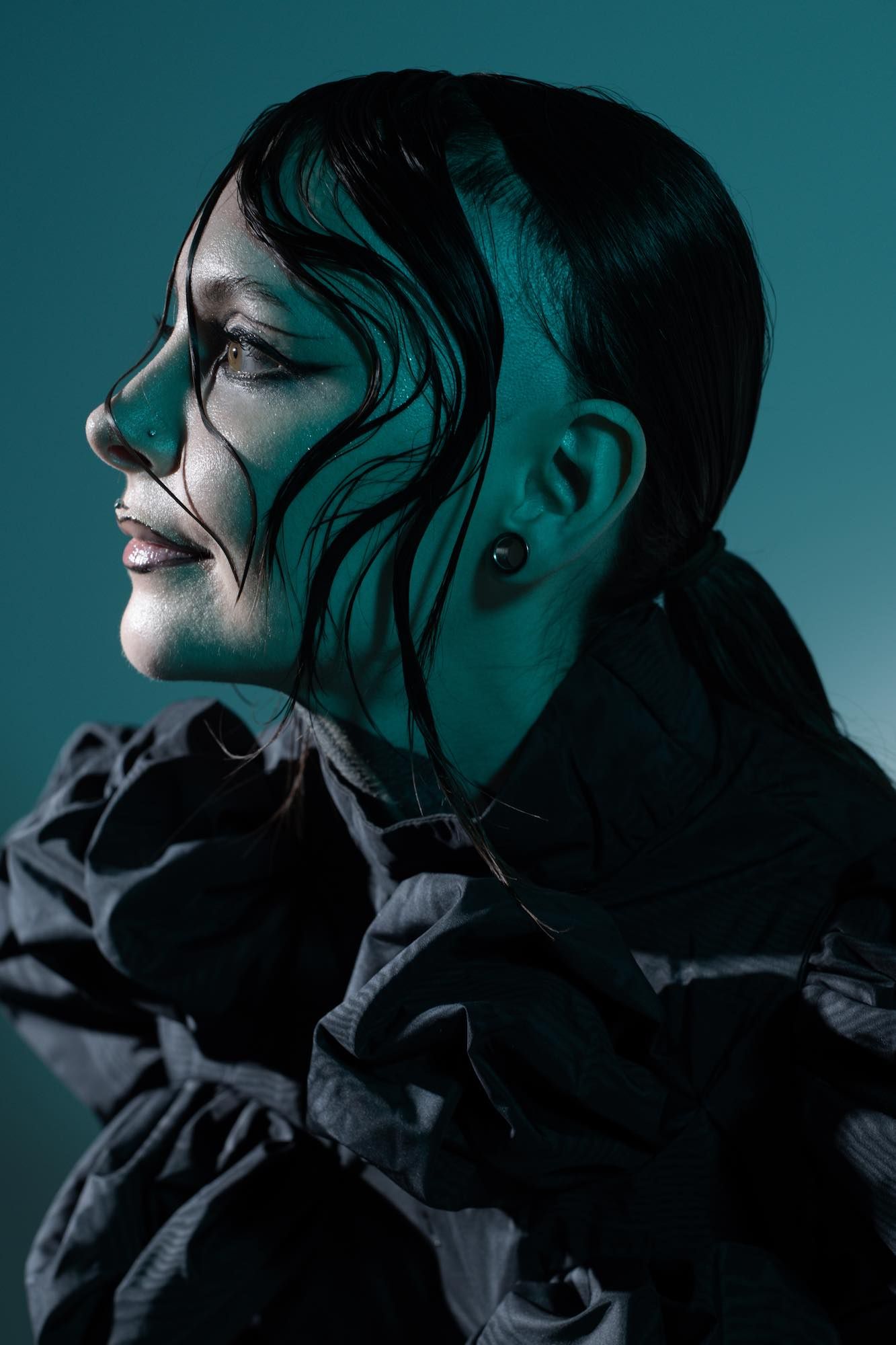 Krista Bourgeois in profile, wearing a ruffled black top under teal studio light