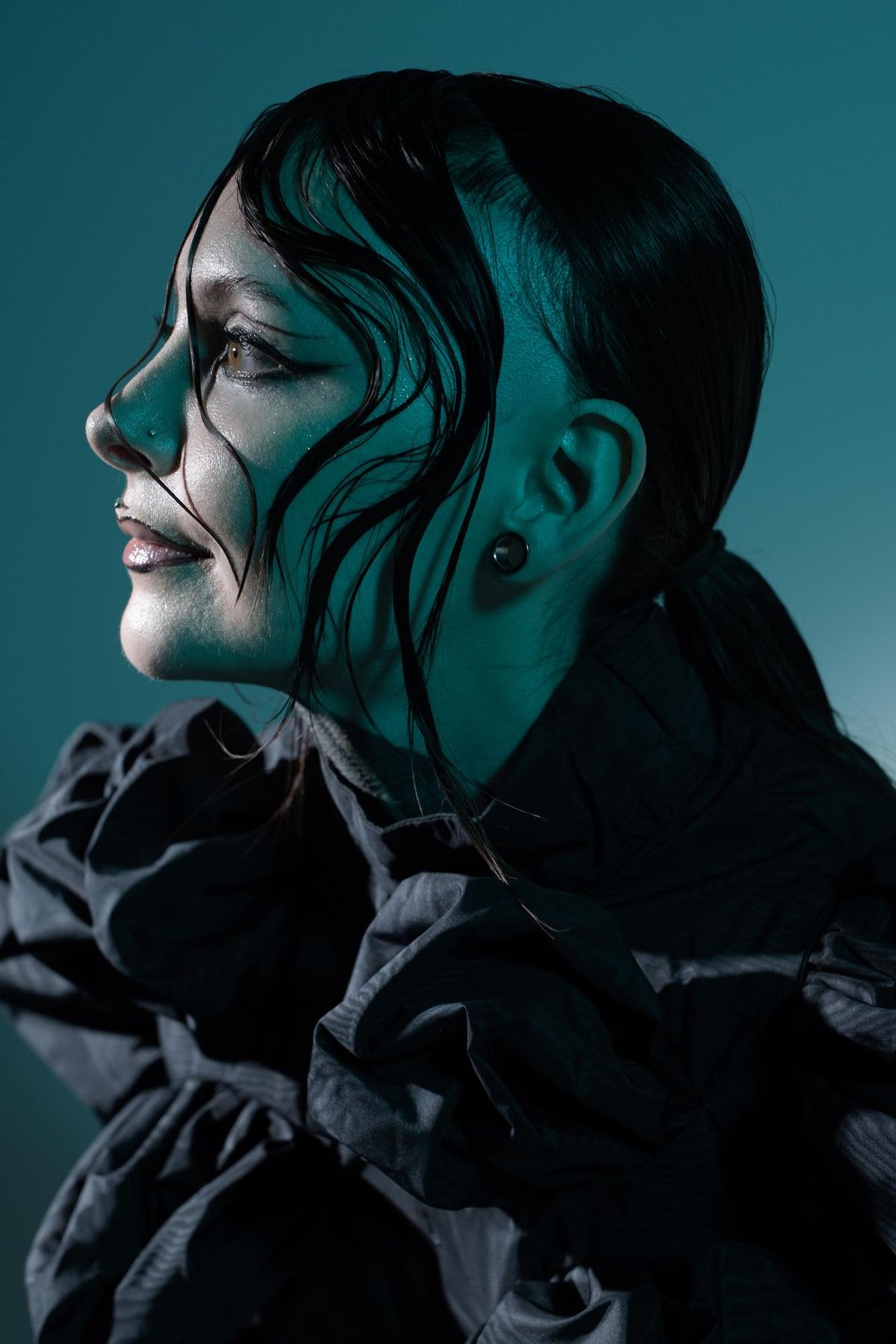 Krista Bourgeois in profile, wearing a ruffled black top under teal studio light