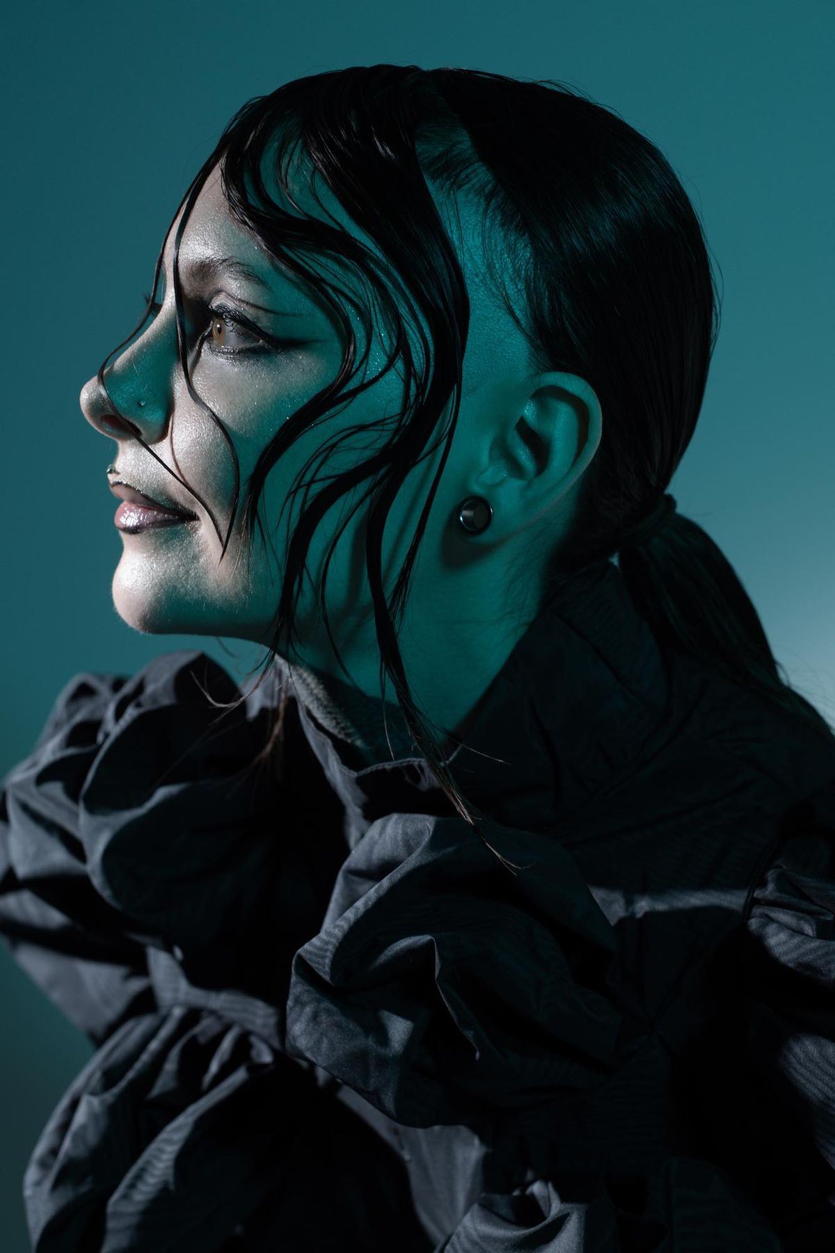 Krista Bourgeois in profile, wearing a ruffled black top under teal studio light