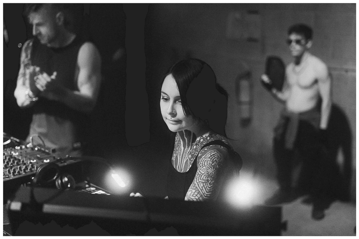 Black-and-white photo of Krista Bourgeois behind a DJ mixer with two people blurred in the background