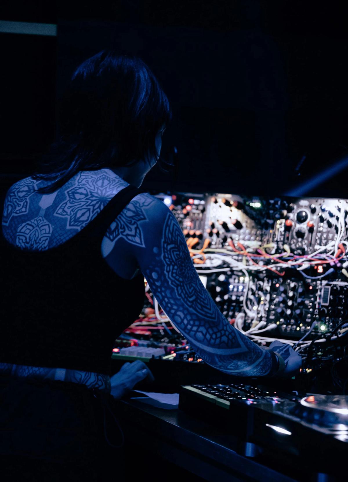 Krista seen from behind, operating a large modular synthesizer in dark blue light
